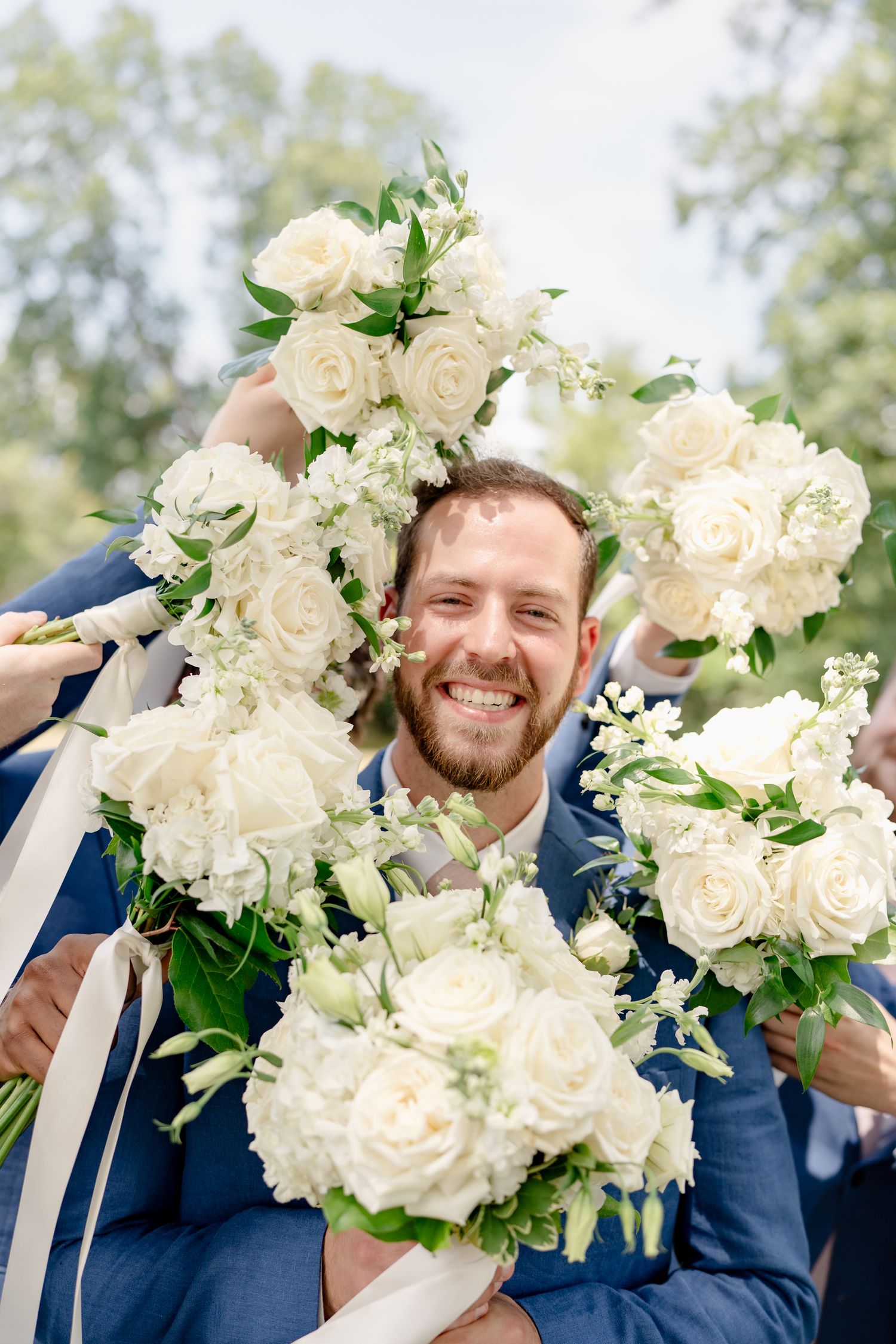 Wedding party members in navy suits hold elegant white rose and floral bouquets during outdoor celebration.