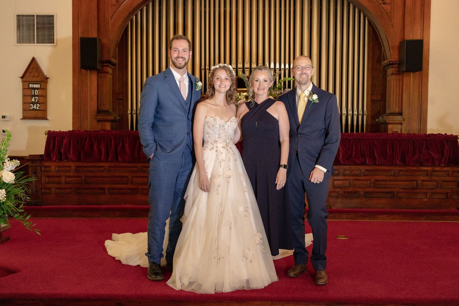 Wedding party members gather on red carpeted church steps in formal attire for group photos.