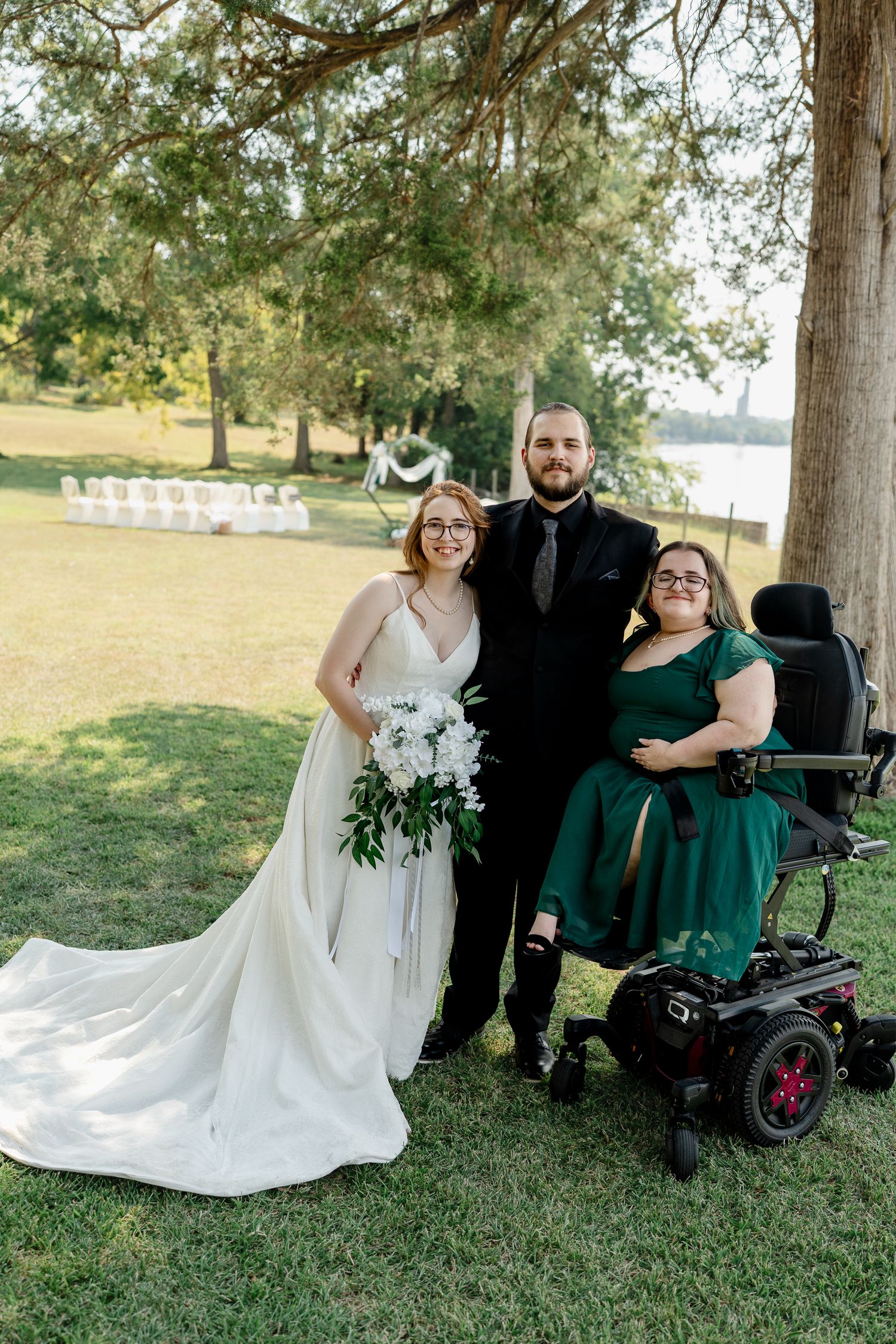 A group in formal attire poses outdoors under trees for wedding photos, with one person seated in a wheelchair.