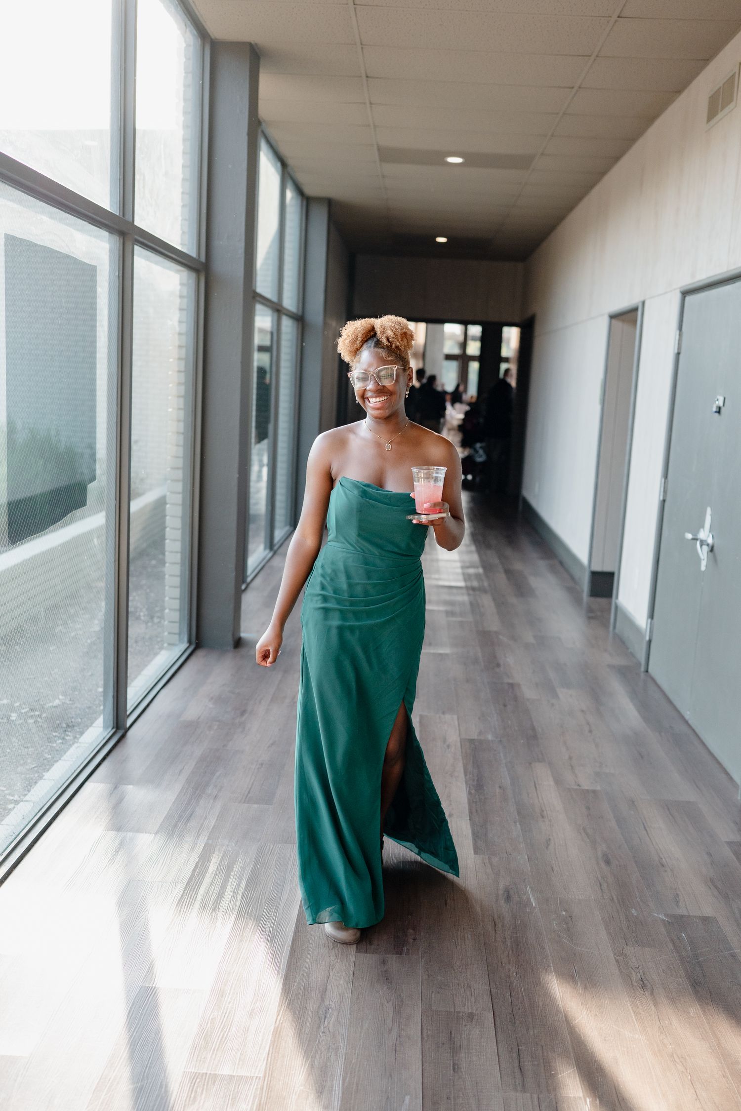 A person in an emerald green formal dress walks down a modern hallway with floor-to-ceiling windows.