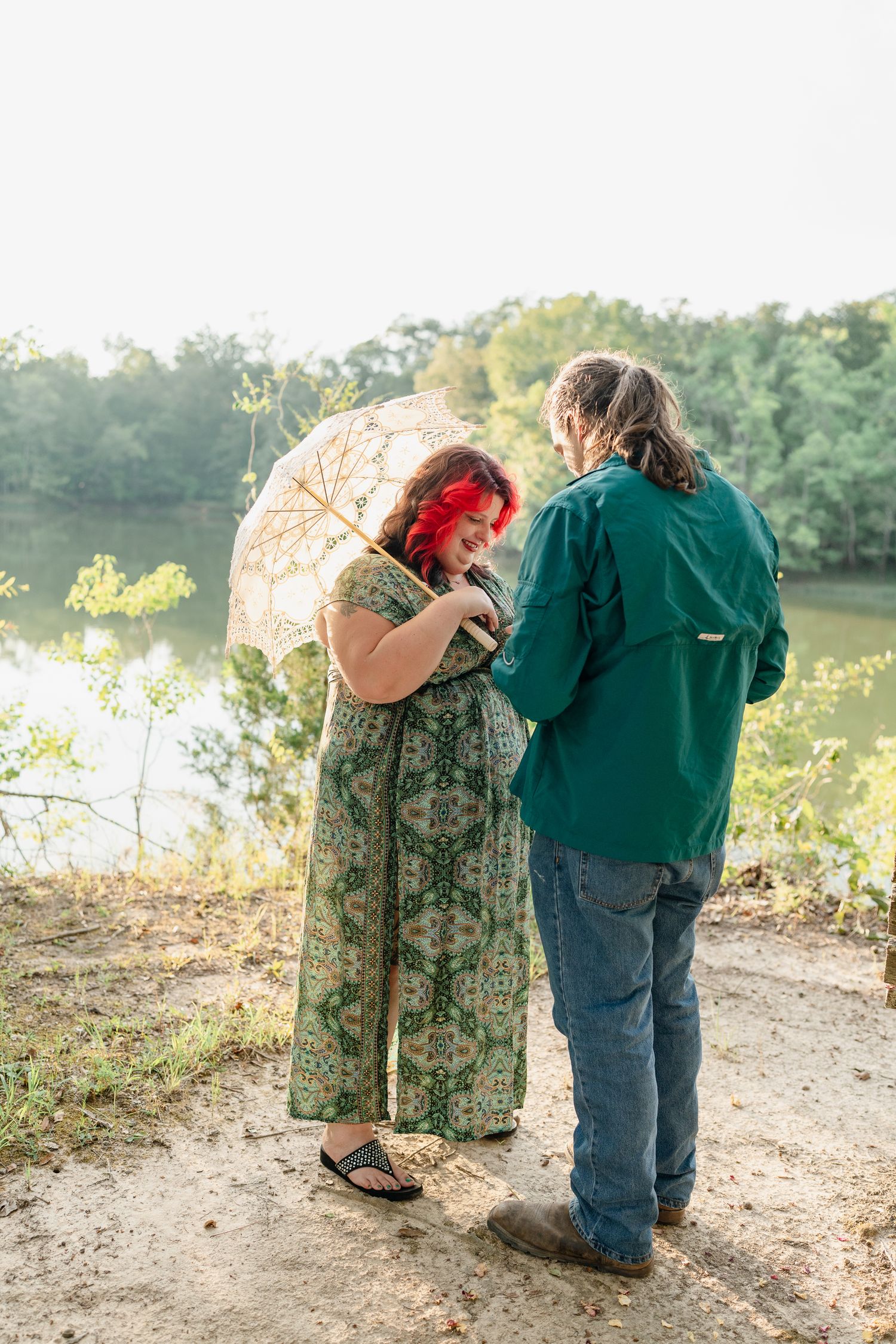 A couple embraces on a dirt path overlooking a lake, with one holding a decorative parasol.