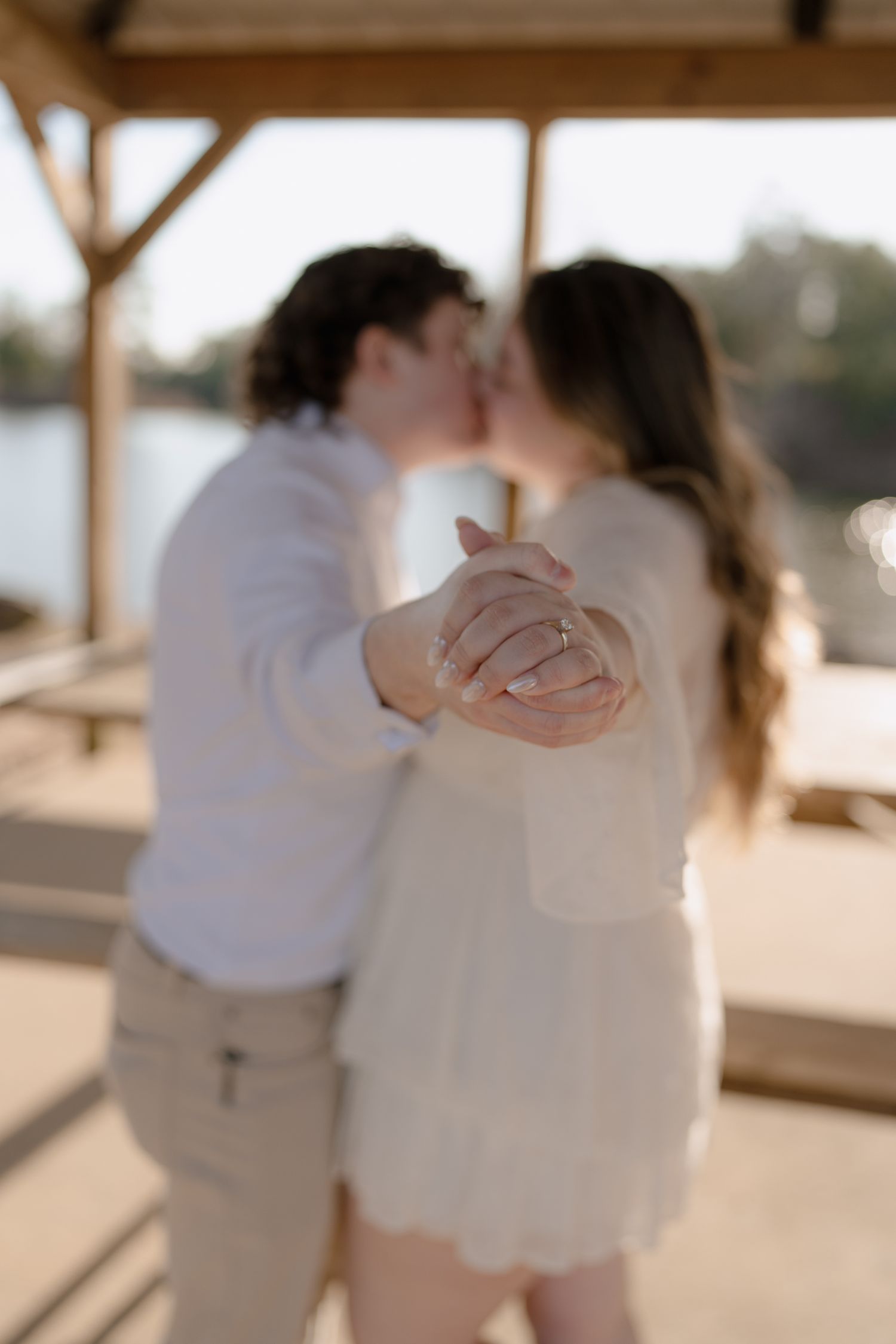Newly engaged couple kissing under wooden gazebo, showcasing engagement ring in soft focus.