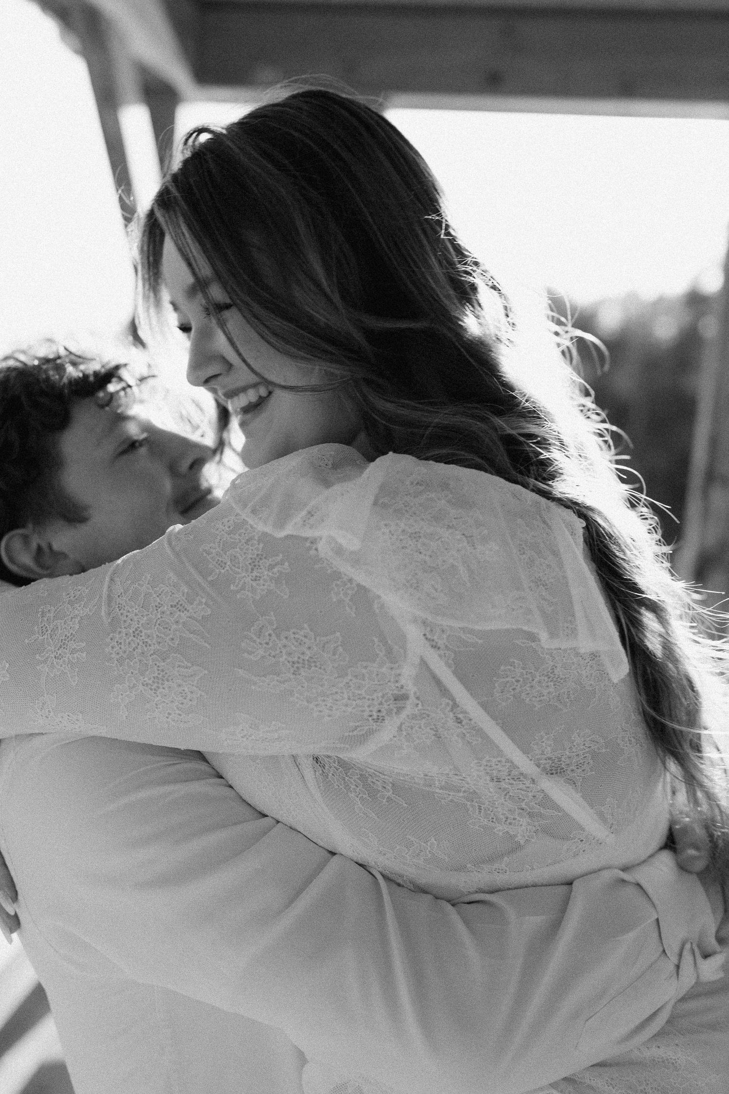 Black and white photo of a smiling woman in lace dress hugging a child outdoors in warm sunlight.