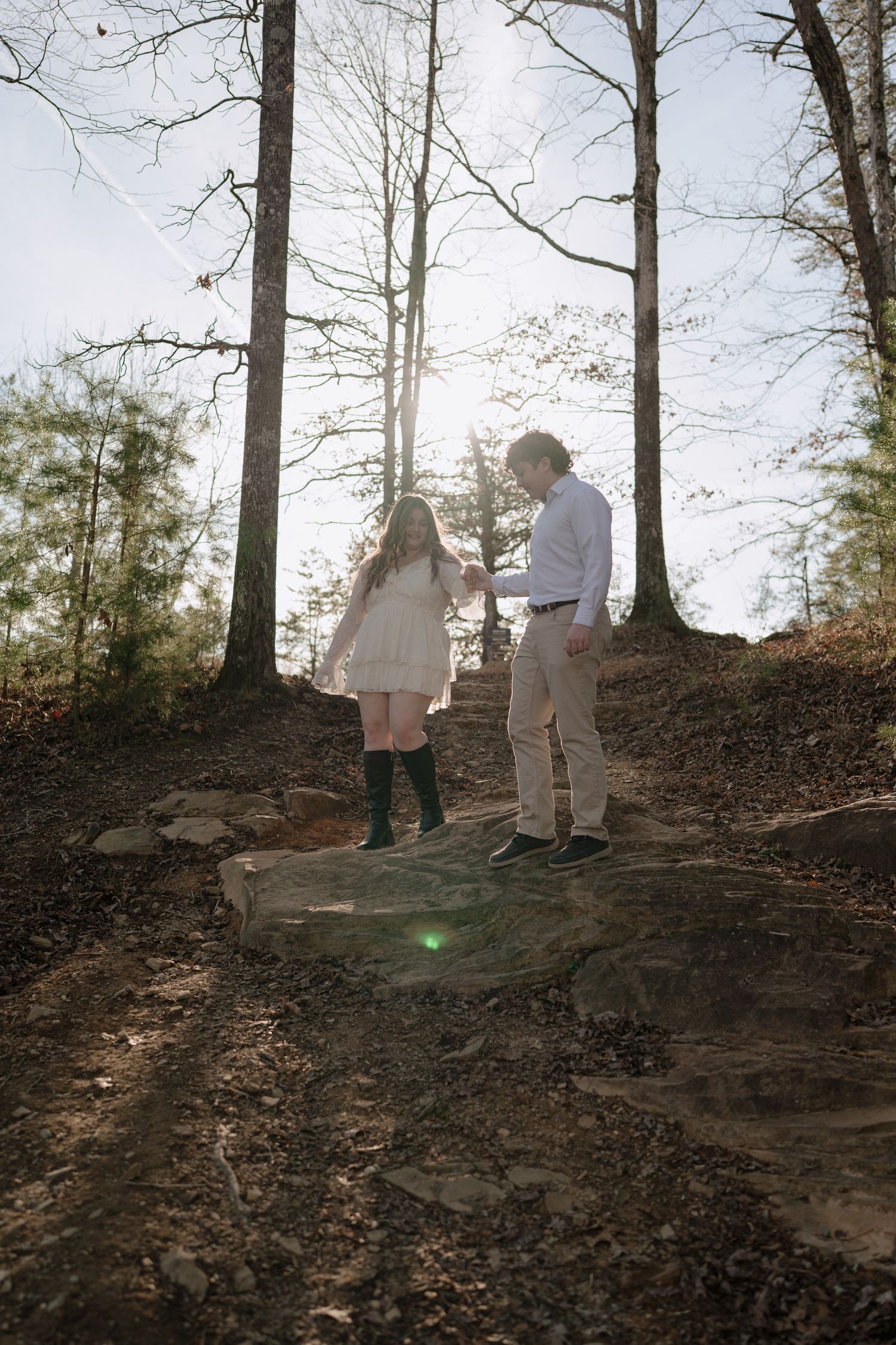 Couple holding hands on rocky forest path with sunlight filtering through bare trees.