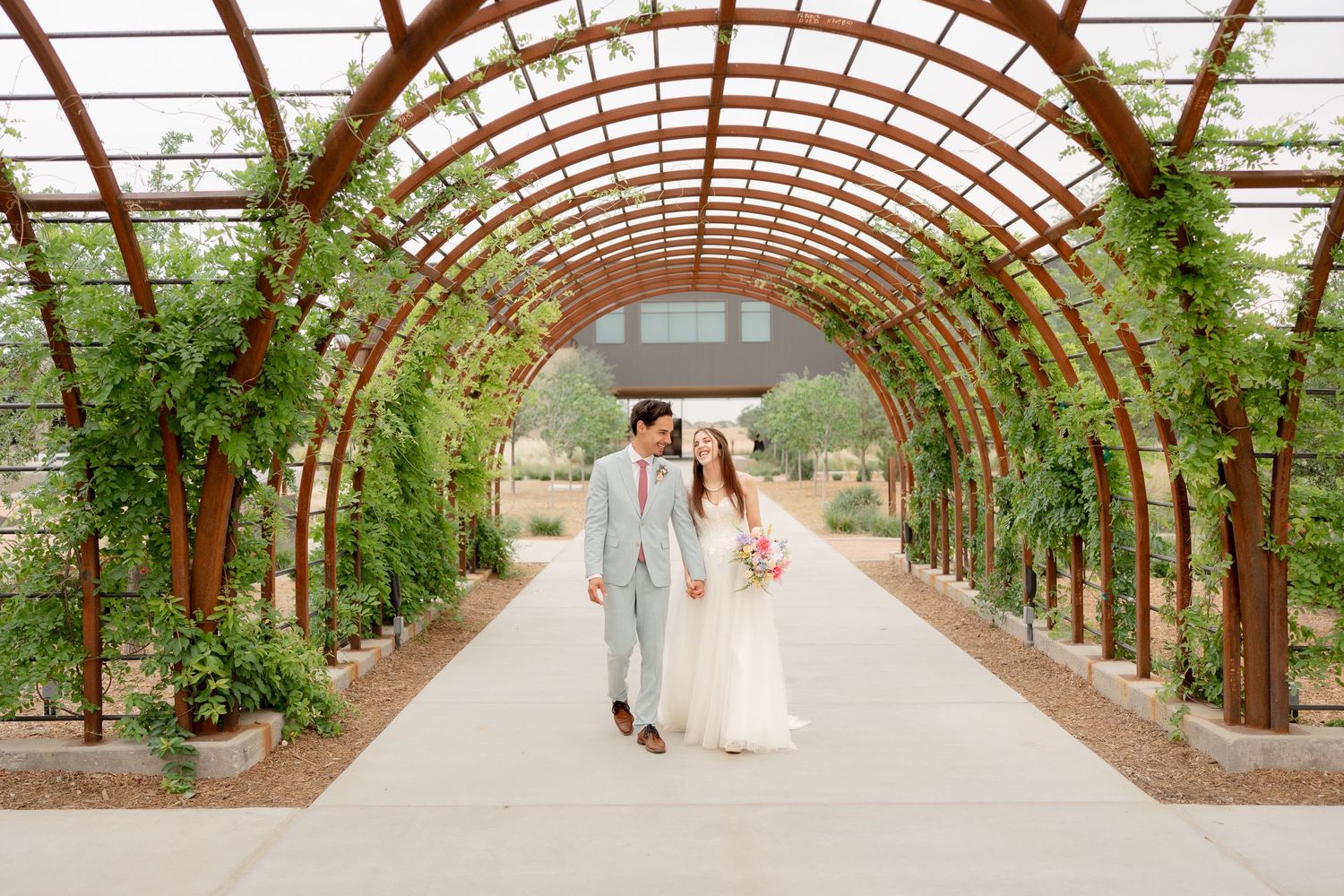 A romantic garden wedding ceremony takes place under a curved wooden pergola covered in lush green vines and flowers.