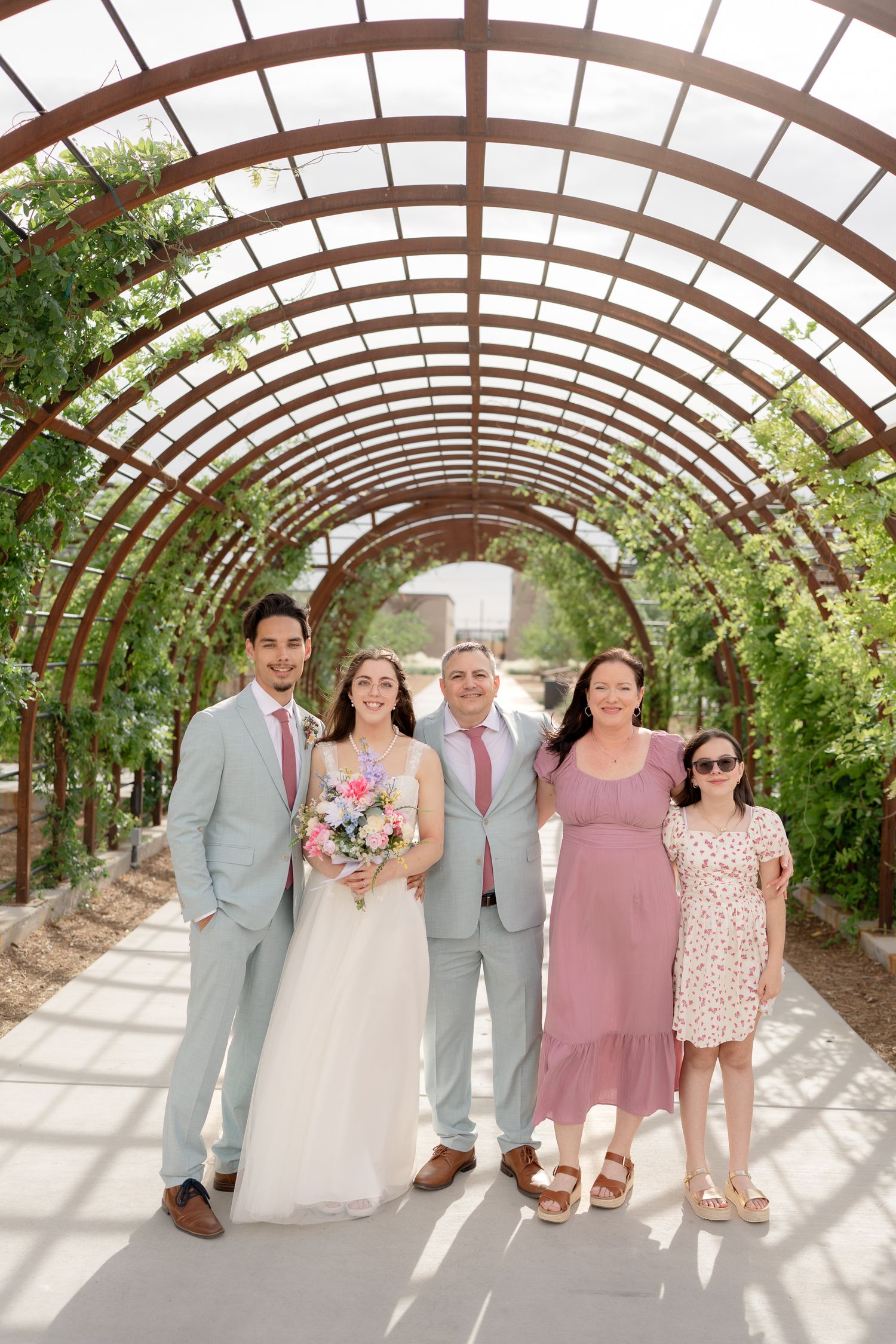 Wedding party poses under curved wooden pergola with greenery in outdoor garden setting.