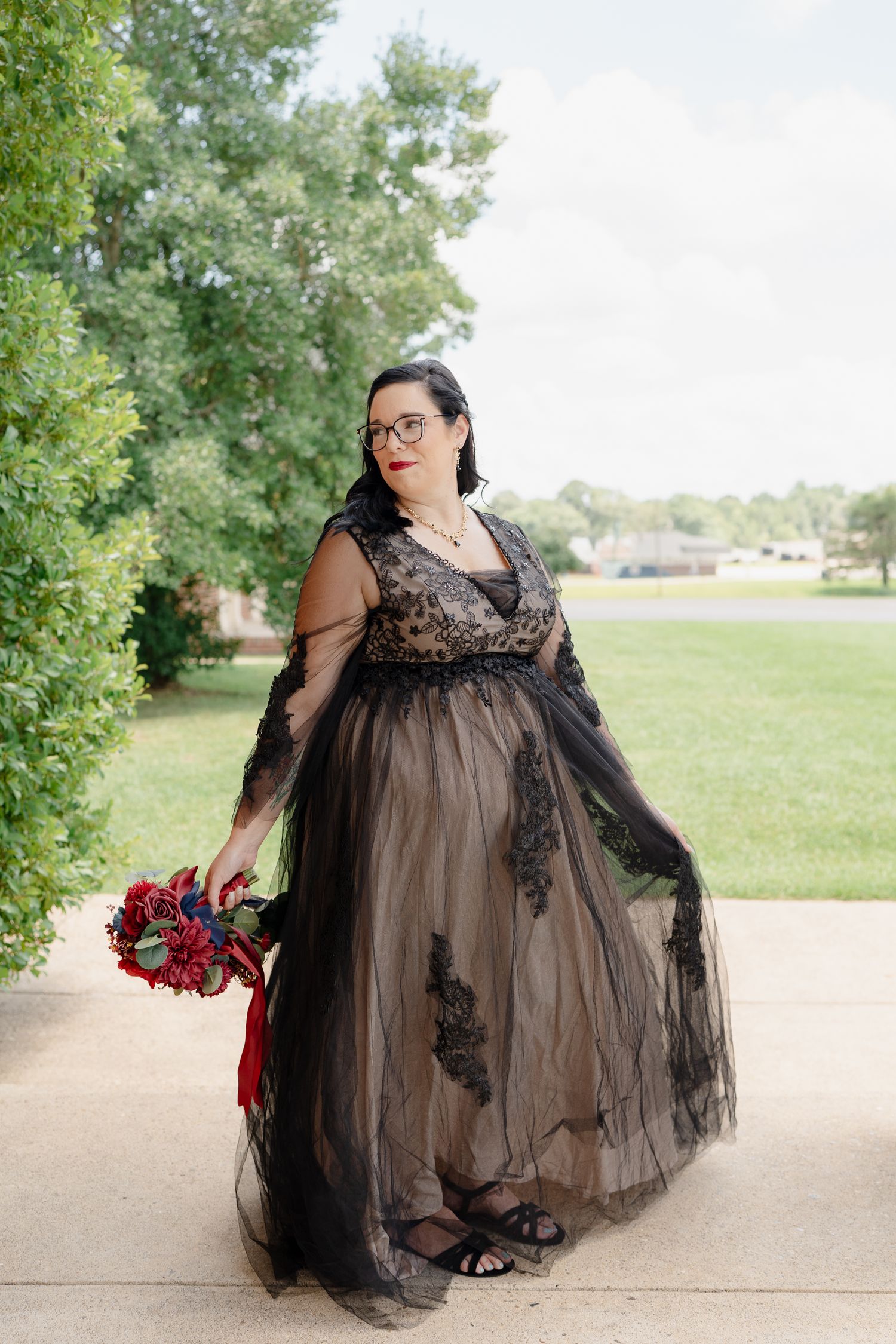 A person in a black lace dress poses with red flowers in an outdoor setting.