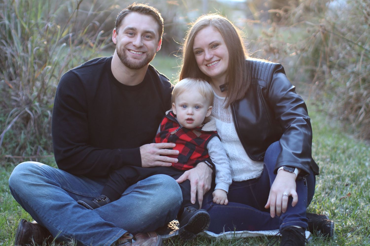 Family outdoor portrait showing parents sitting with toddler in red plaid vest on grass.