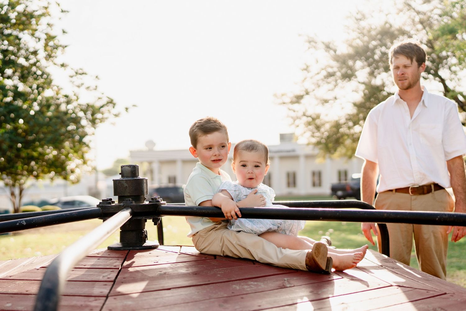 Children sit on a red surface while an adult stands nearby during a sunny outdoor photoshoot.