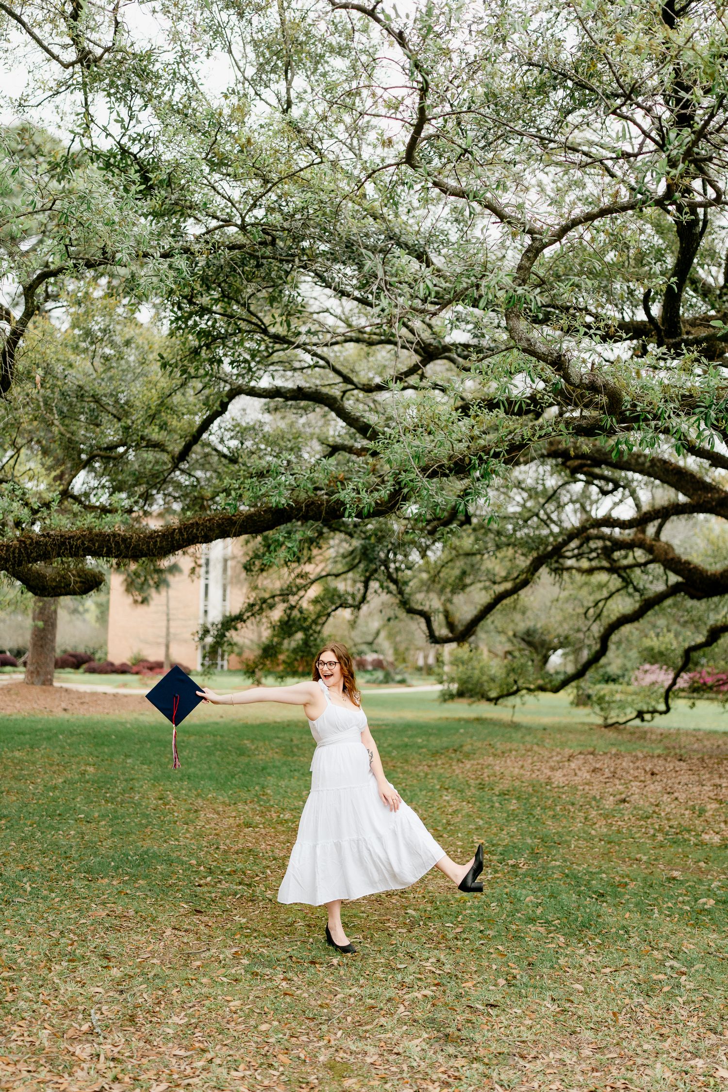 Graduate twirls in white dress under canopy of oak trees during photo session.