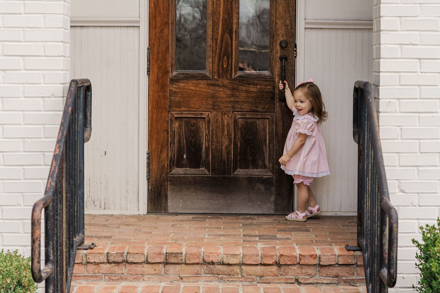 Small child in pink dress stands on brick steps in front of wooden door.