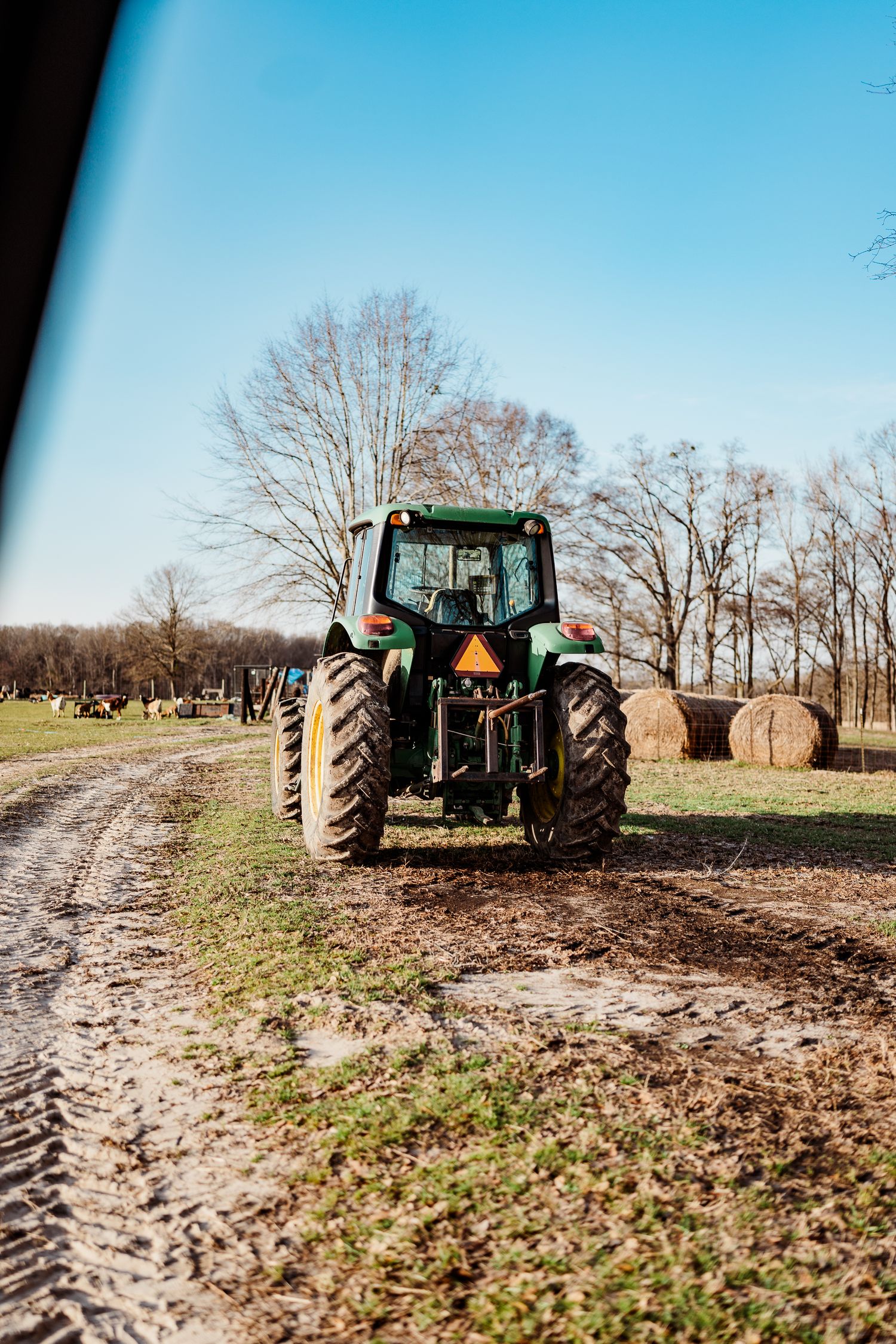 Green tractor working in a muddy field during winter with bare trees in background.