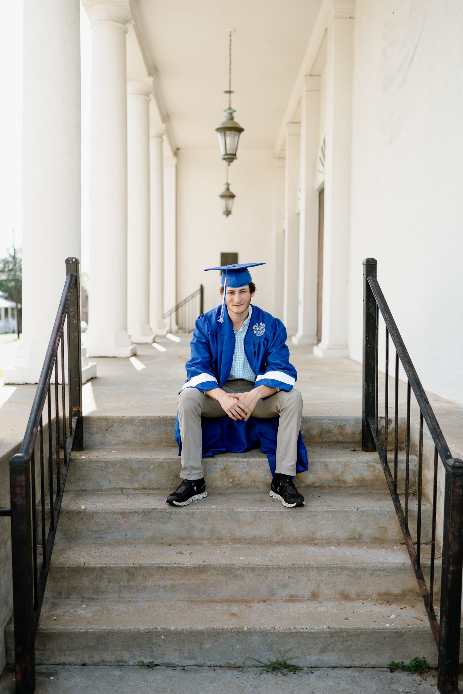 Graduate in blue regalia sits on concrete steps between elegant white columns.