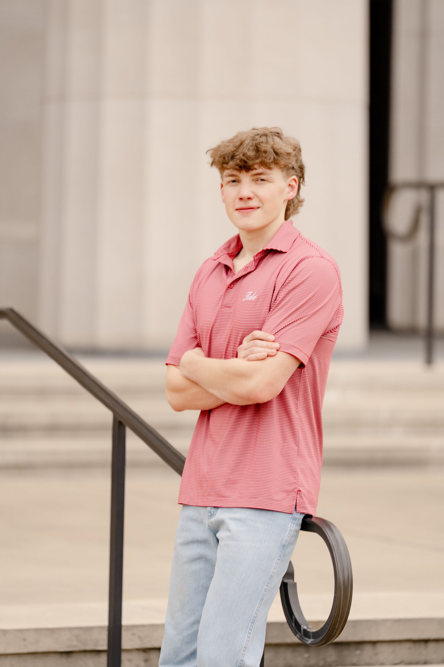 Person in a pink polo and jeans poses casually by stone steps with arms crossed.