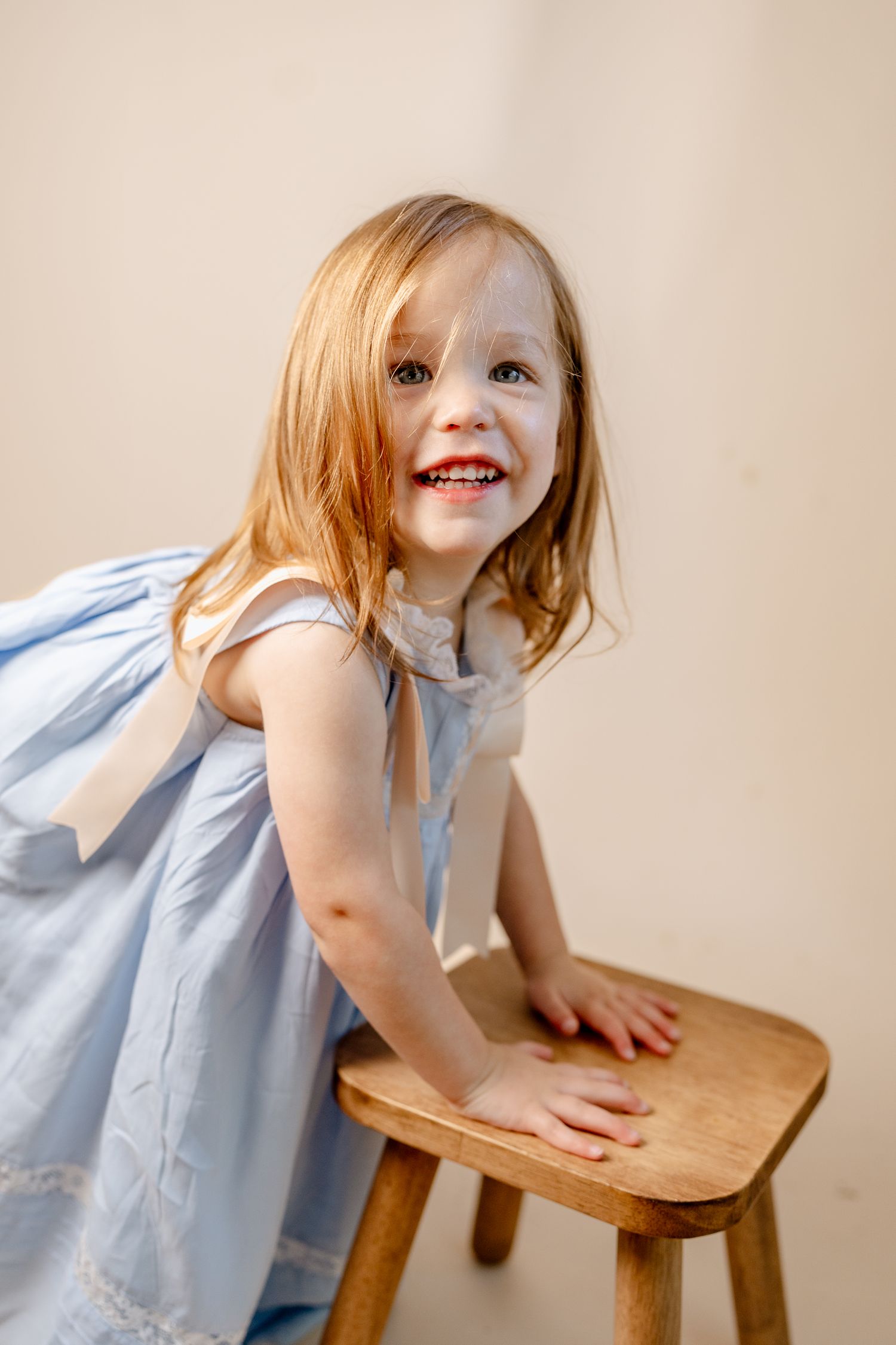 A child in a light blue dress plays on a wooden stool against a neutral background.