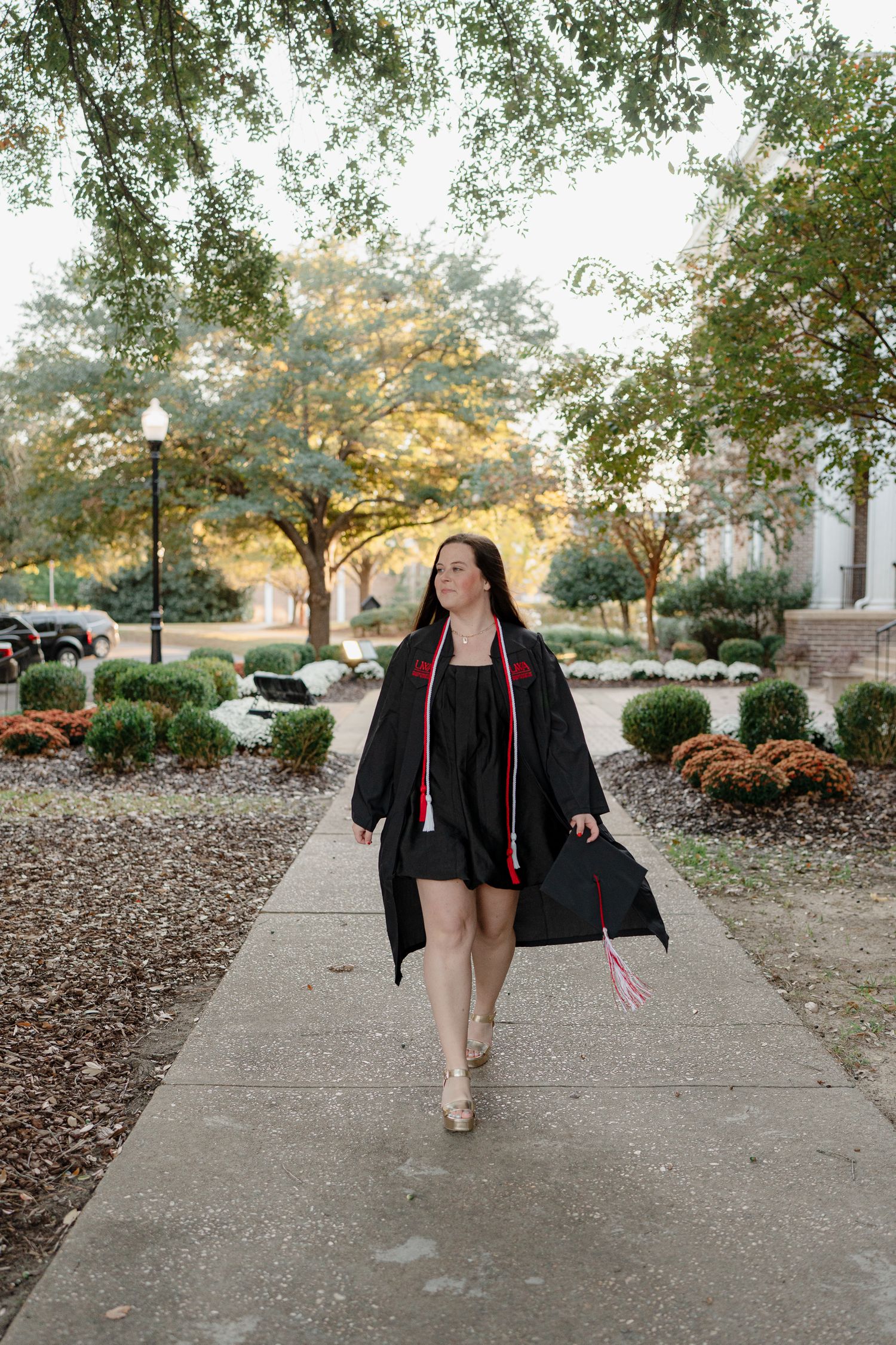 Graduate walking on concrete path surrounded by landscaped grounds and trees in autumn.