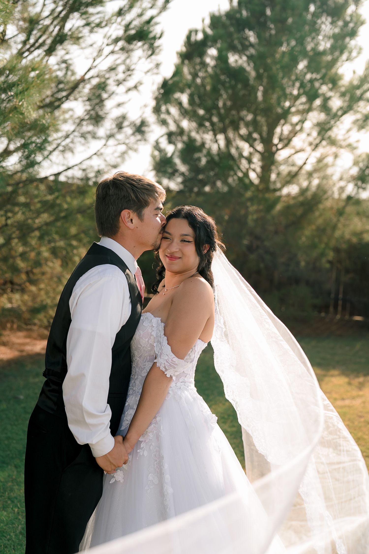Bride smiles as groom kisses her cheek outdoors at golden hour, her veil flowing in the breeze.