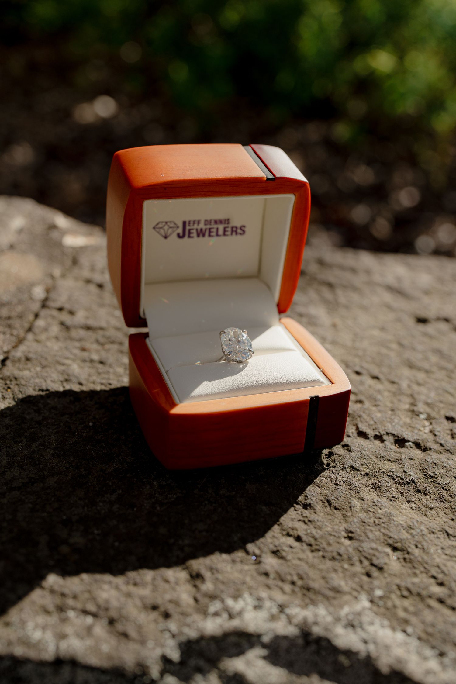 A diamond engagement ring displayed in an orange Jeff Bennie Jewelers box resting on a stone surface.