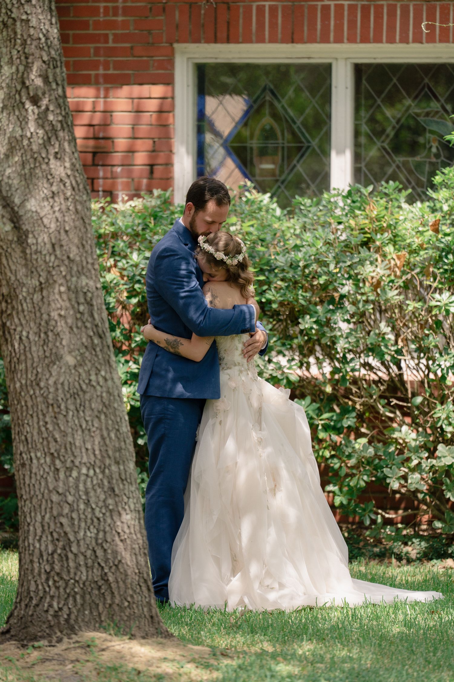 A romantic first look moment between newlyweds in front of a brick building with flowering bushes and trees.