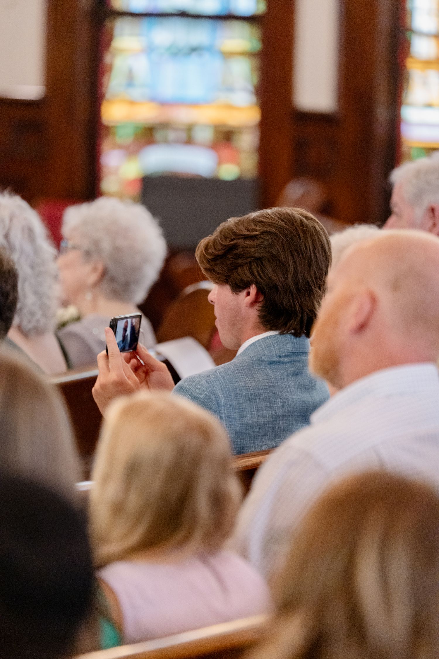 A congregation sits in wooden pews inside a church with stained glass windows visible in the background.