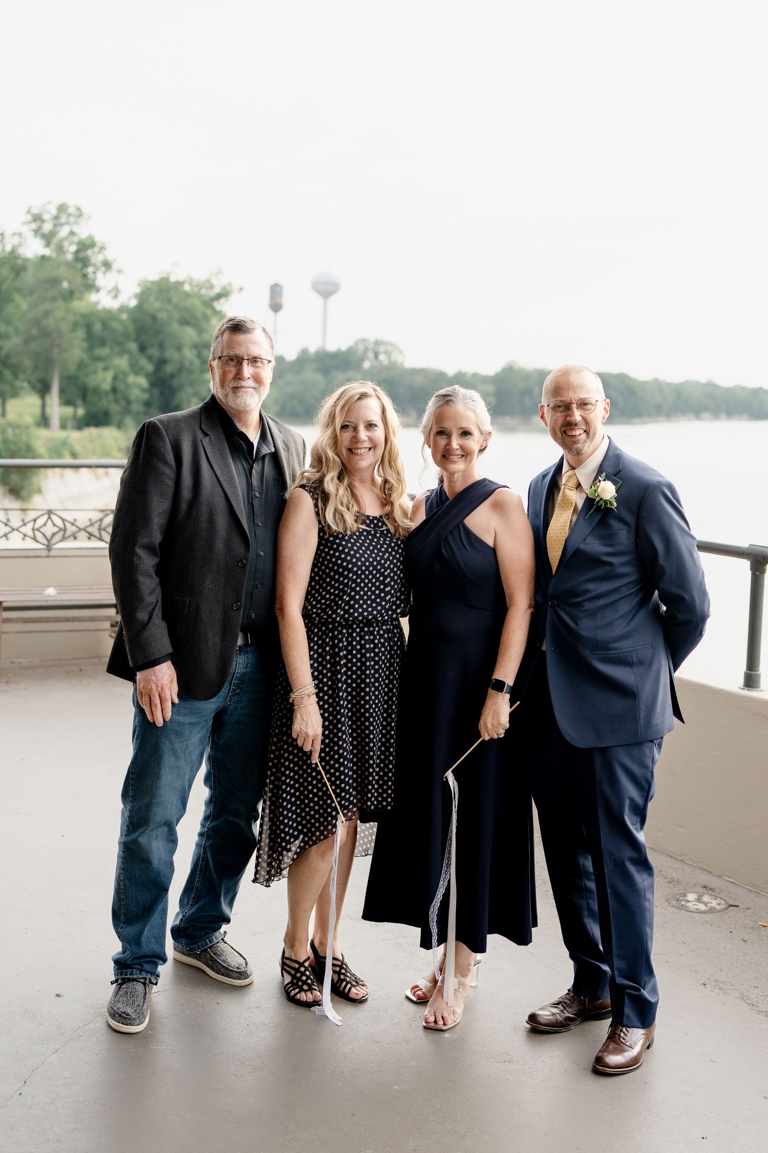 Four people in formal attire pose together on a covered outdoor patio with water views in the background.