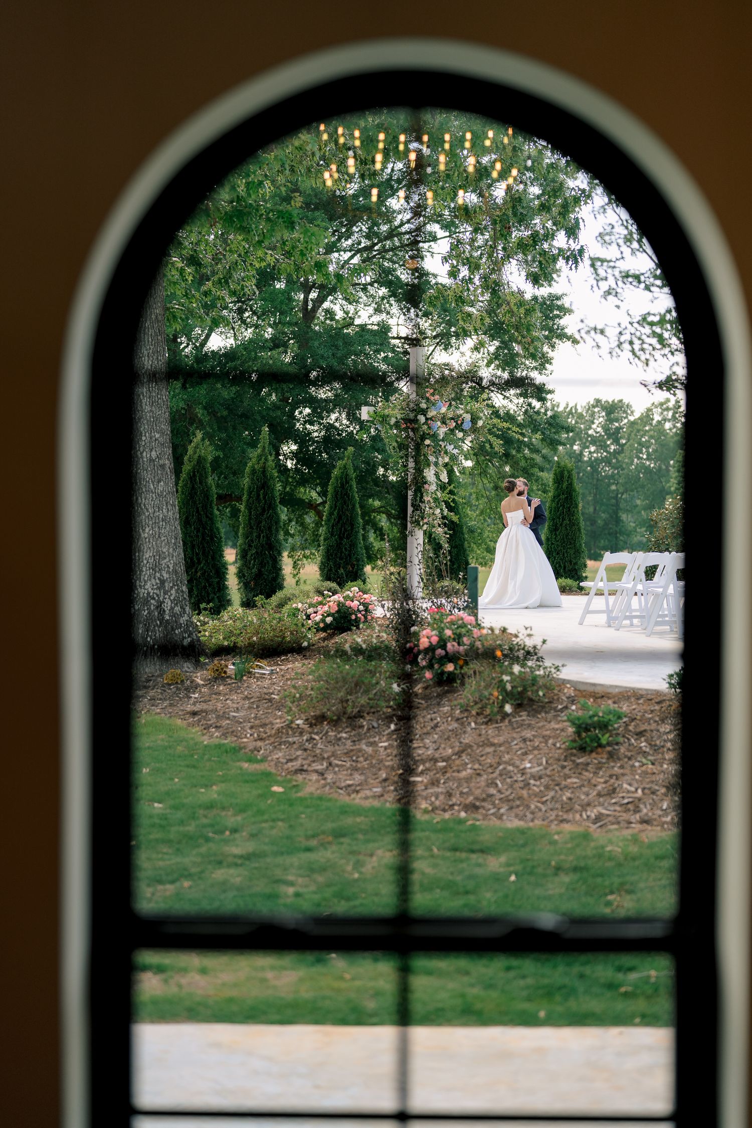 Bride in white gown viewed through arched window, surrounded by lush gardens and elegant outdoor wedding chairs.