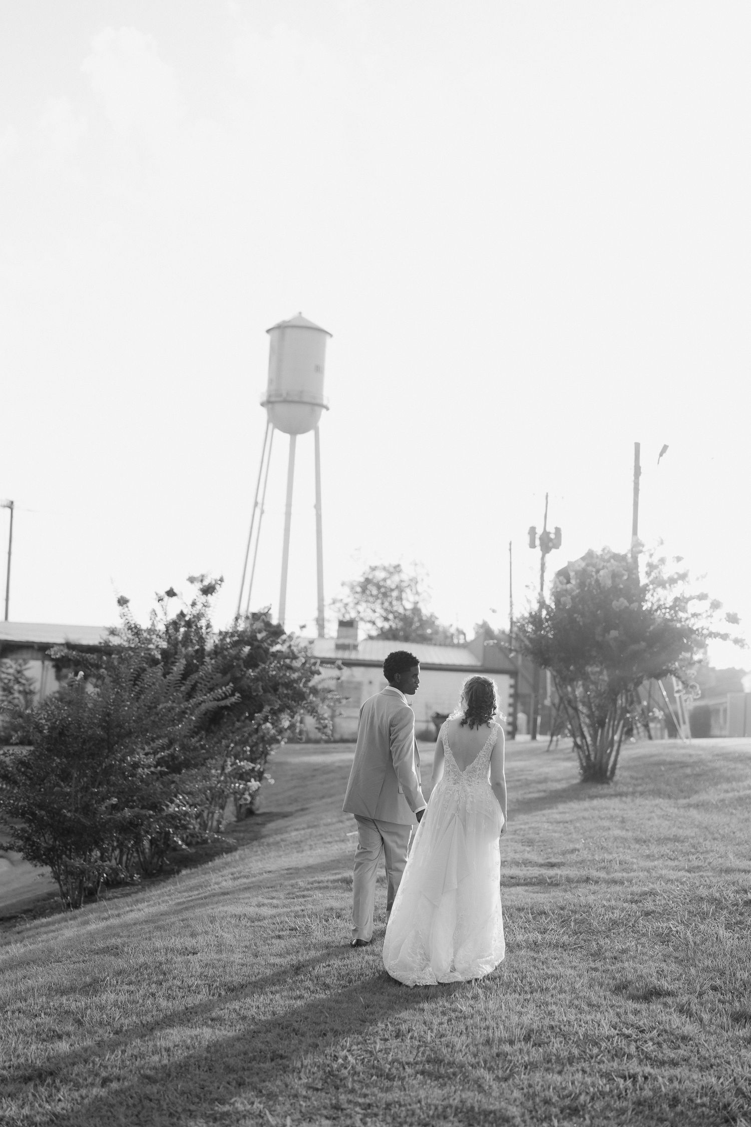 Black and white photo of newlyweds walking hand in hand near a water tower at sunset.