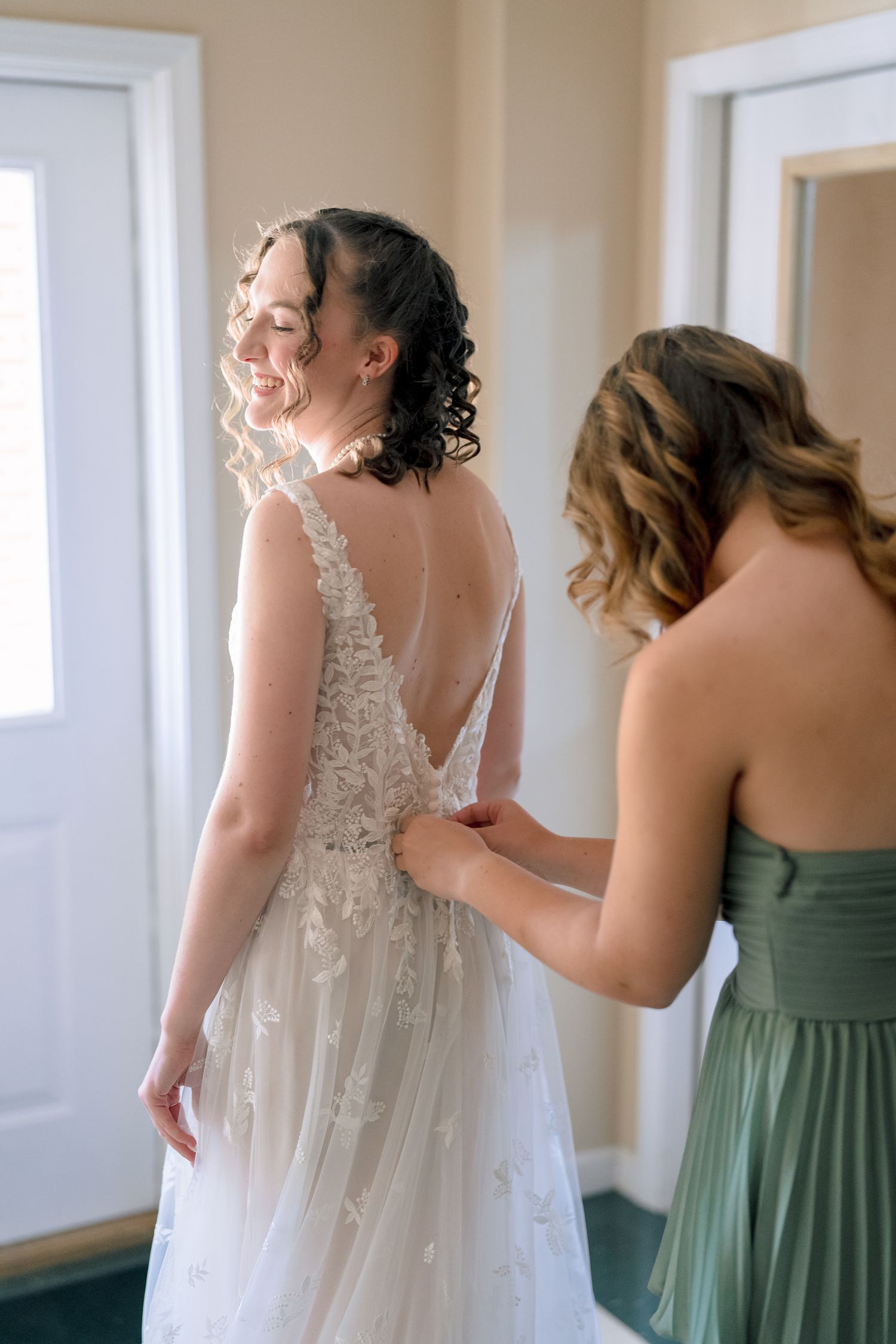A bridesmaid in sage green helps a smiling bride in a lace backless wedding gown get dressed before the ceremony.