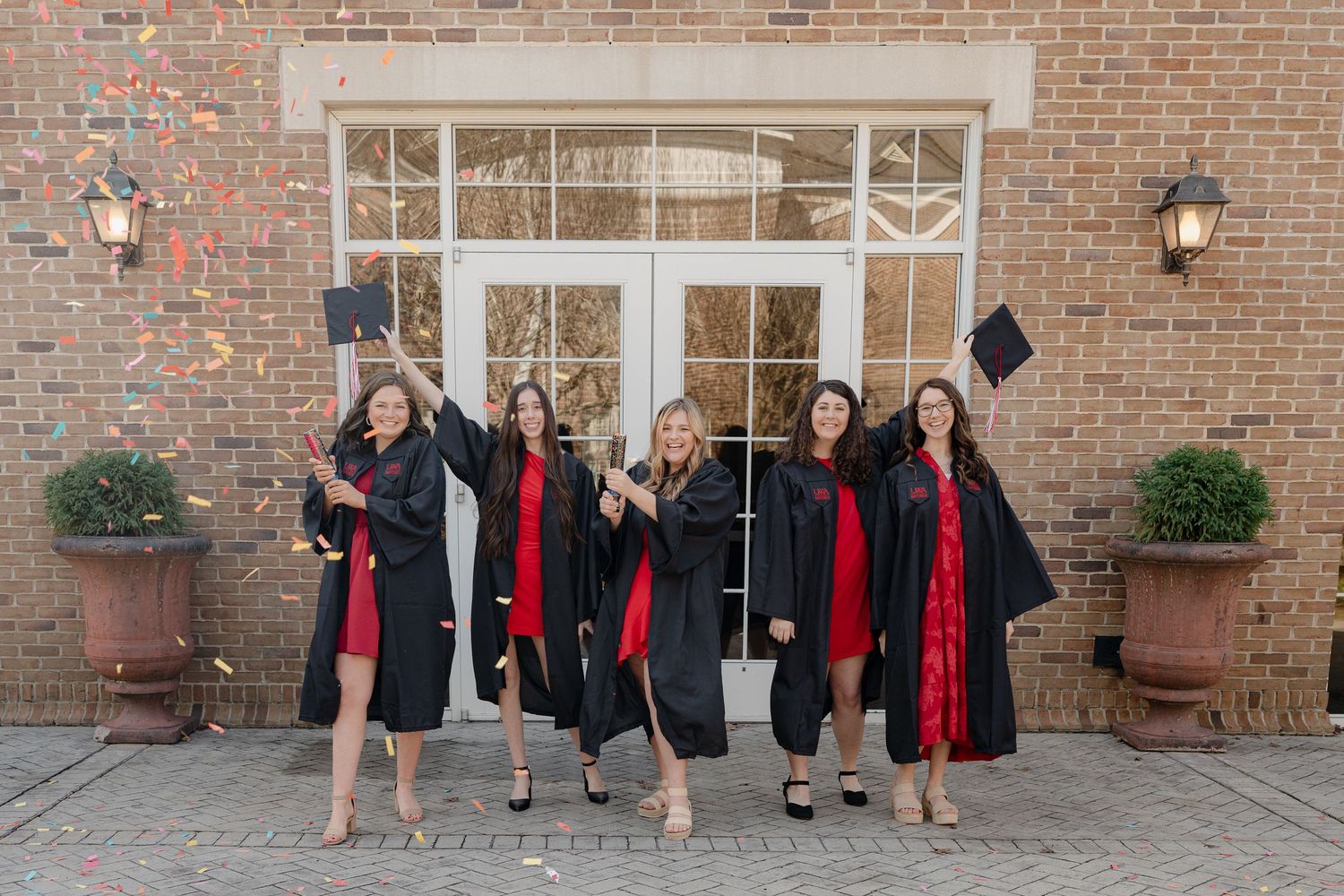 Graduates in black caps and gowns with red stoles celebrate together outside brick building entrance.