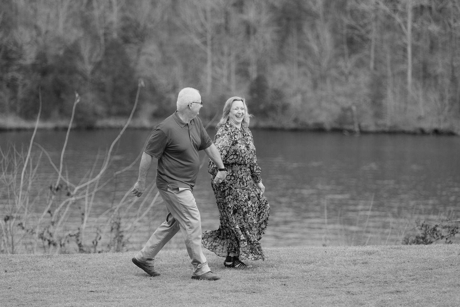 Black and white photo of a couple walking hand in hand along a lakeside path surrounded by trees.