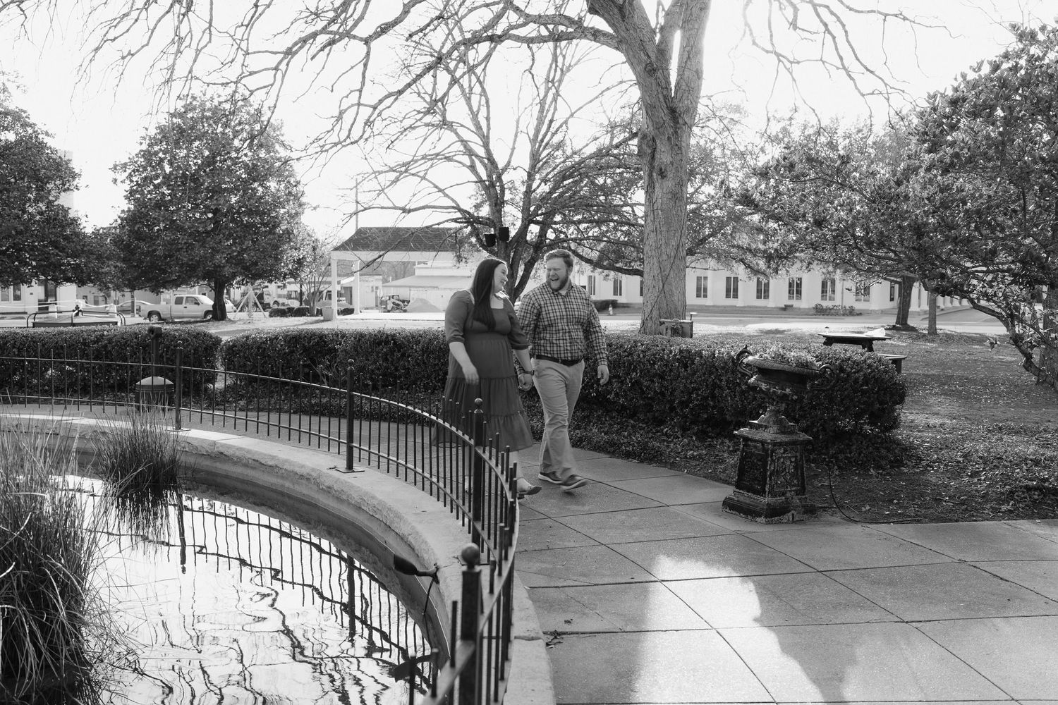 Black and white photo of a couple strolling along a curved garden path beside a small pond with trees.
