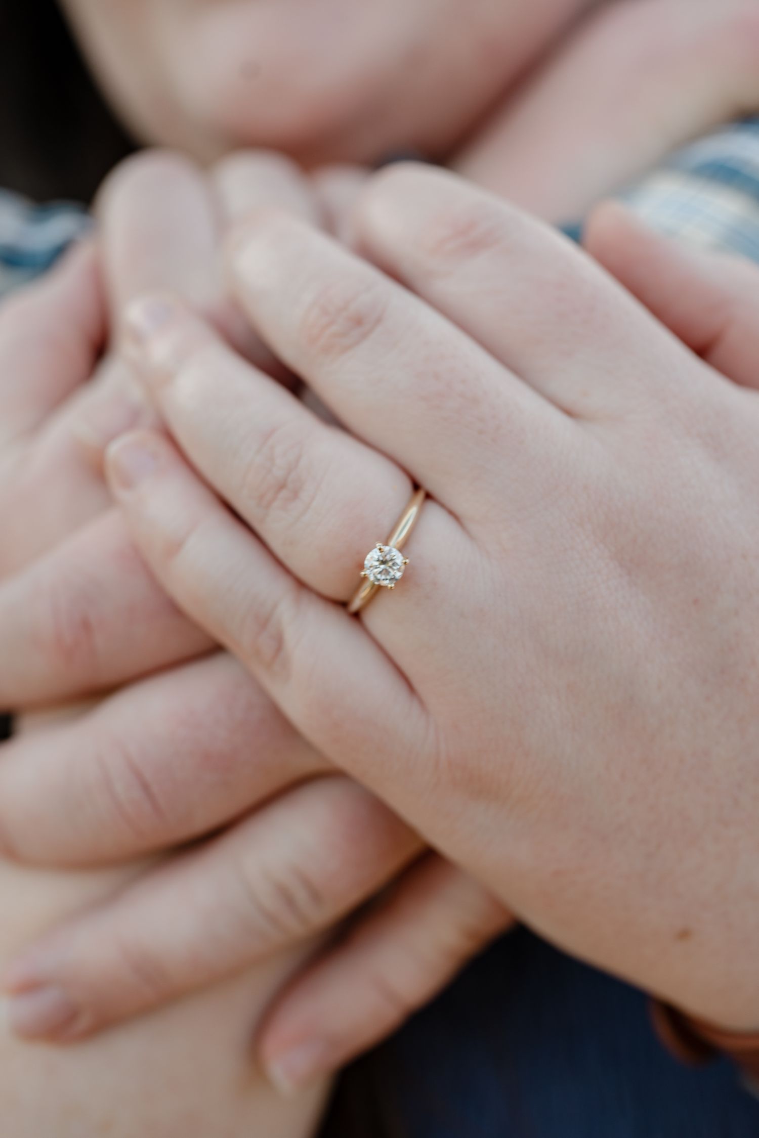 Close-up of hands wearing a delicate gold solitaire diamond engagement ring, with two people embracing.