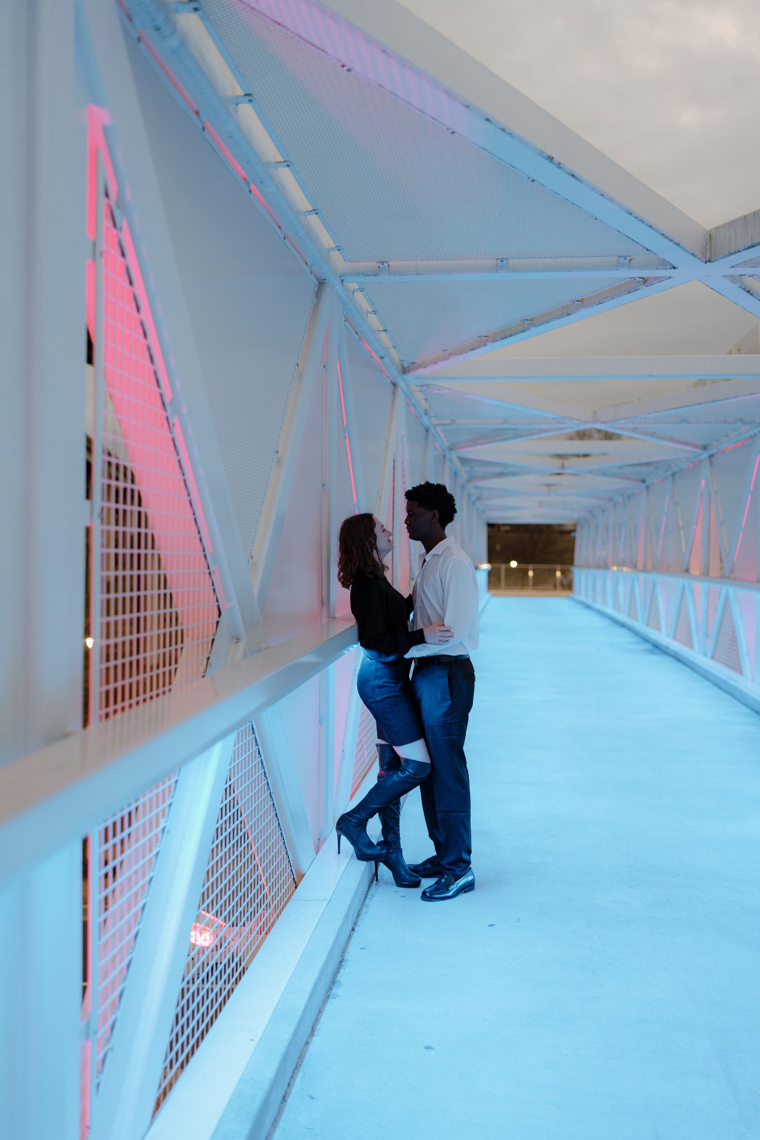 Interracial couple sharing an intimate moment leaning against the railing of a modern bridge with pink and blue lighting.