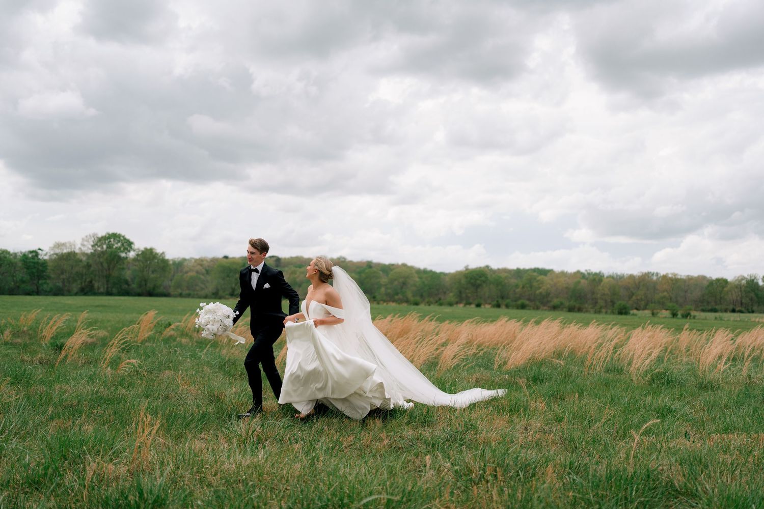 A bride and groom walk hand-in-hand through a lush green field on their wedding day under a cloudy sky.