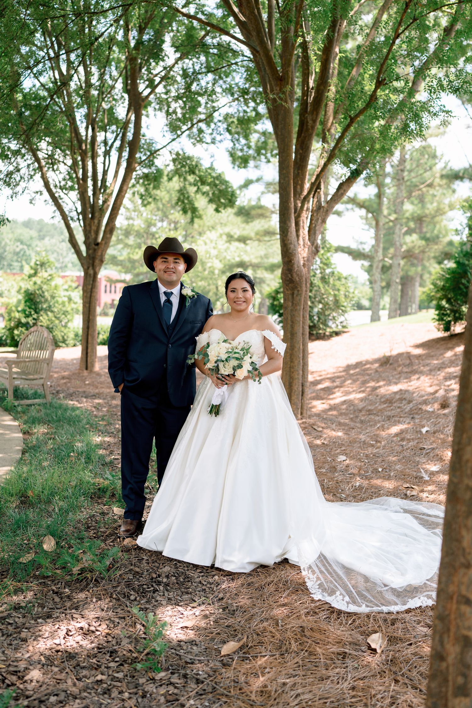 A wedding couple poses under tall trees on a wooded path, with the bride in a flowing white gown holding a bouquet.
