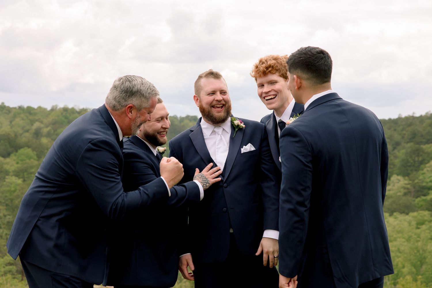 Groom with red beard sharing joyful moment with groomsmen in navy suits outdoors.