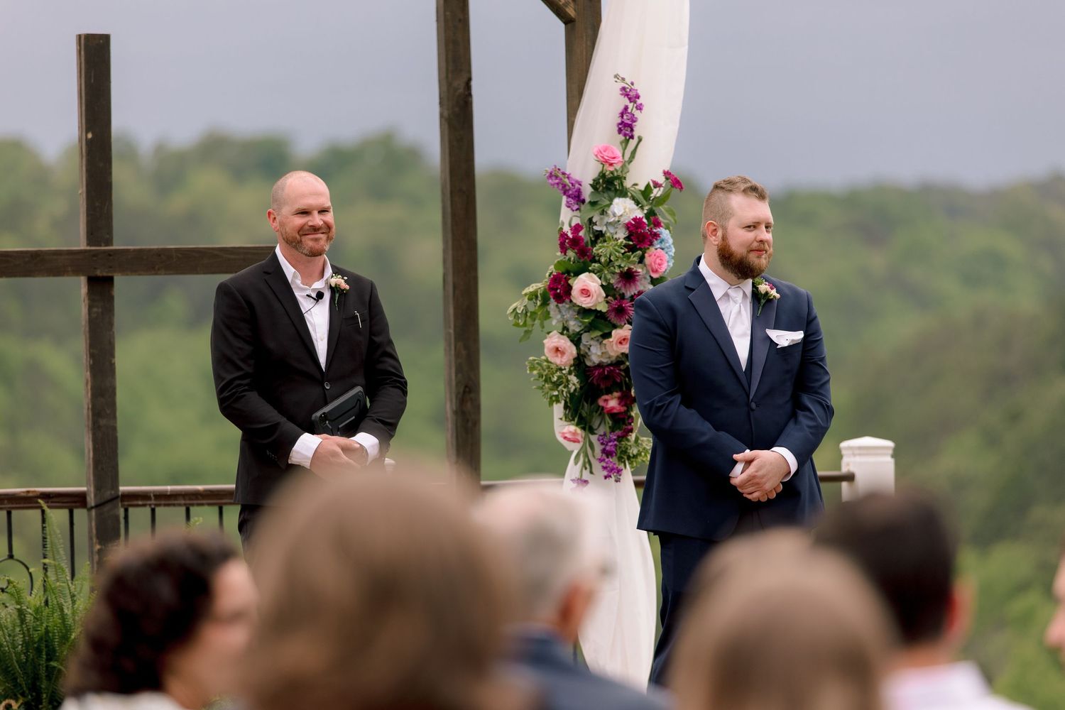 Two men in suits stand at an outdoor wedding altar adorned with colorful floral arrangements.