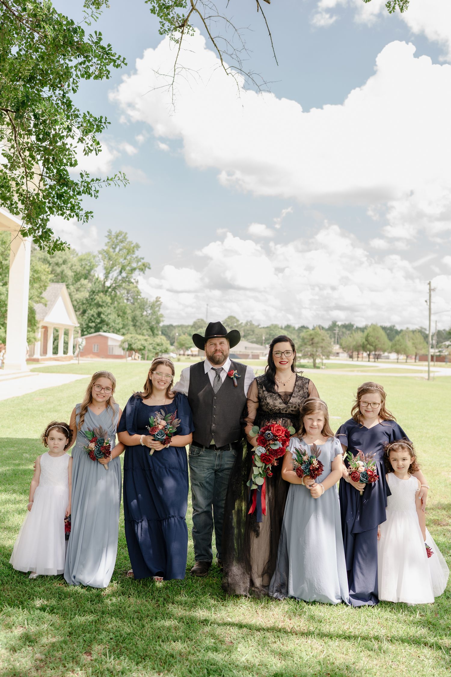A wedding party lineup poses outdoors on a grassy lawn with trees and blue sky in the background.