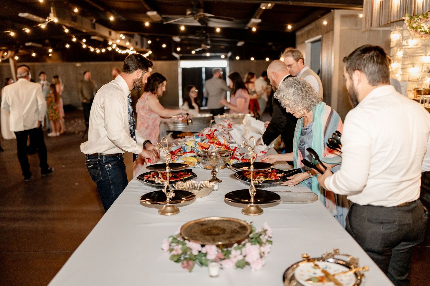 Guests gather around elegantly decorated dining tables under warm string lights at an indoor event venue.
