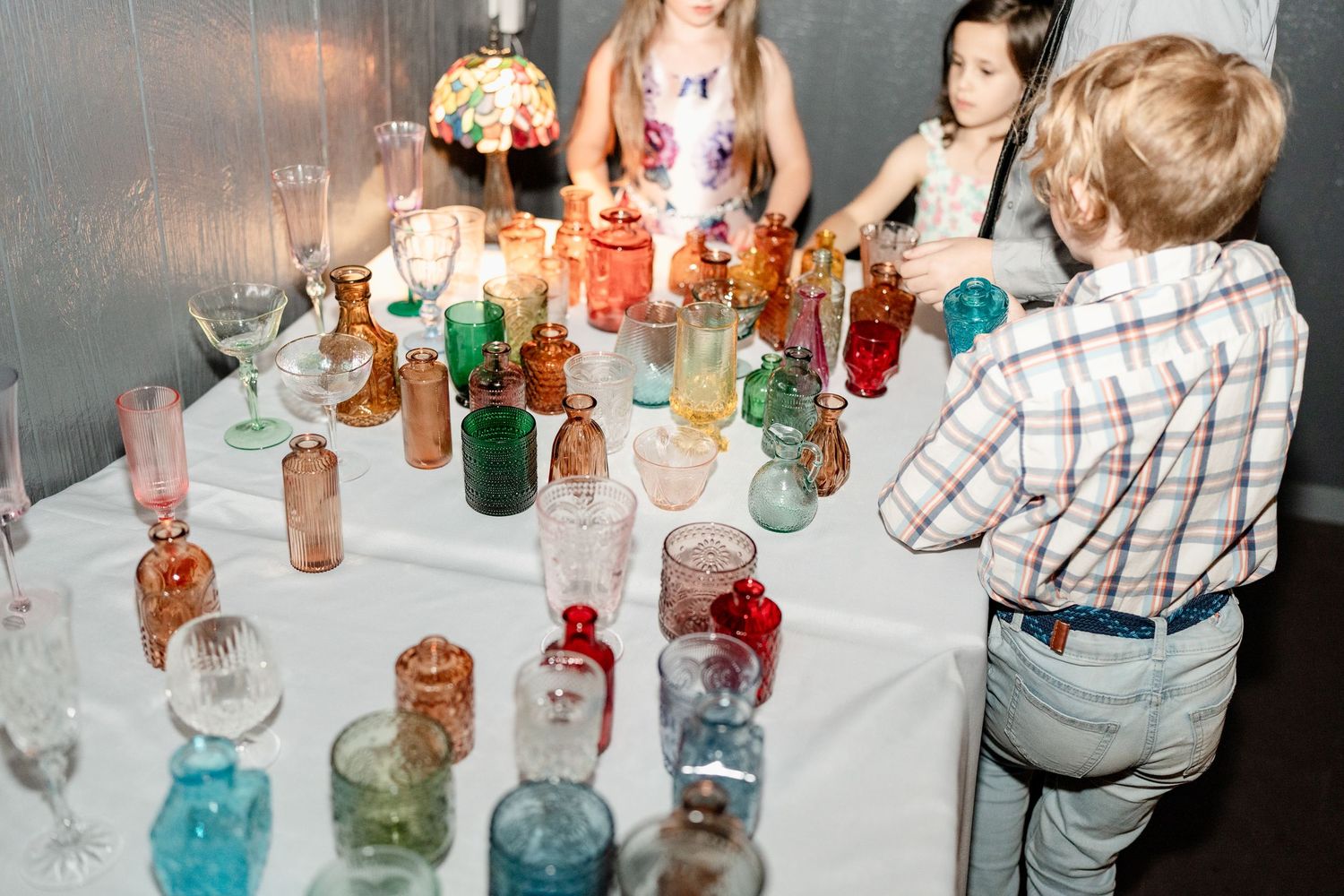 People gathered around a table displaying a collection of colorful vintage glass bottles and jars in various shapes and sizes.