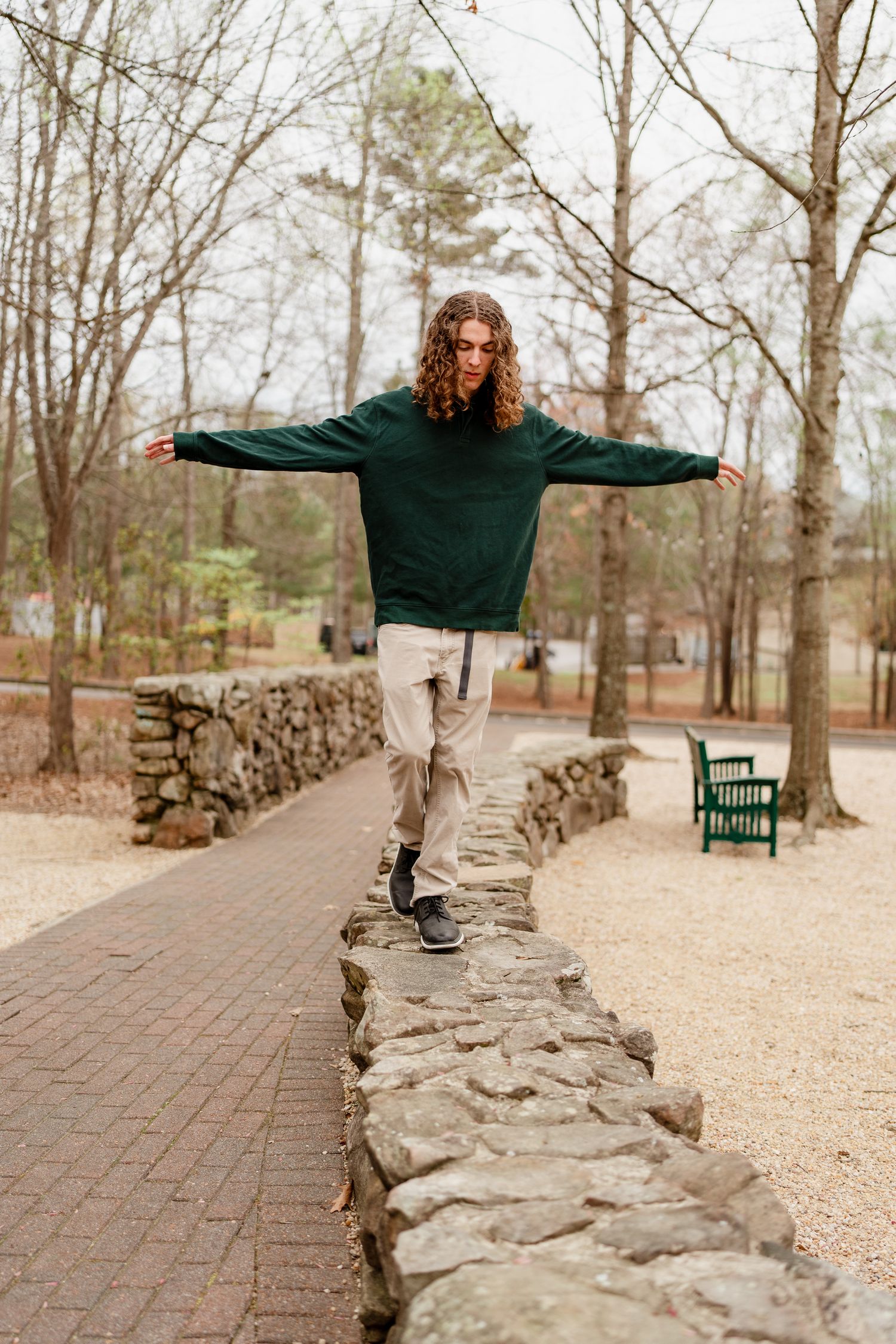 Figure in green sweater balancing with arms outstretched on stone wall in park setting.