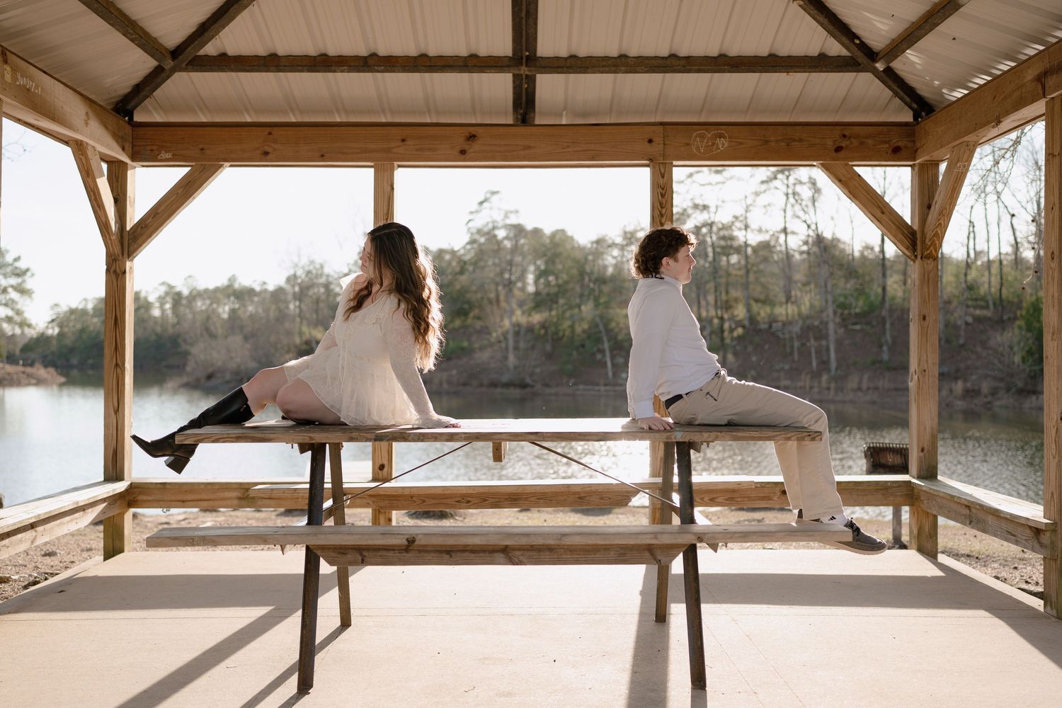 Couple sitting back-to-back on a picnic table bench inside a wooden lakeside gazebo.