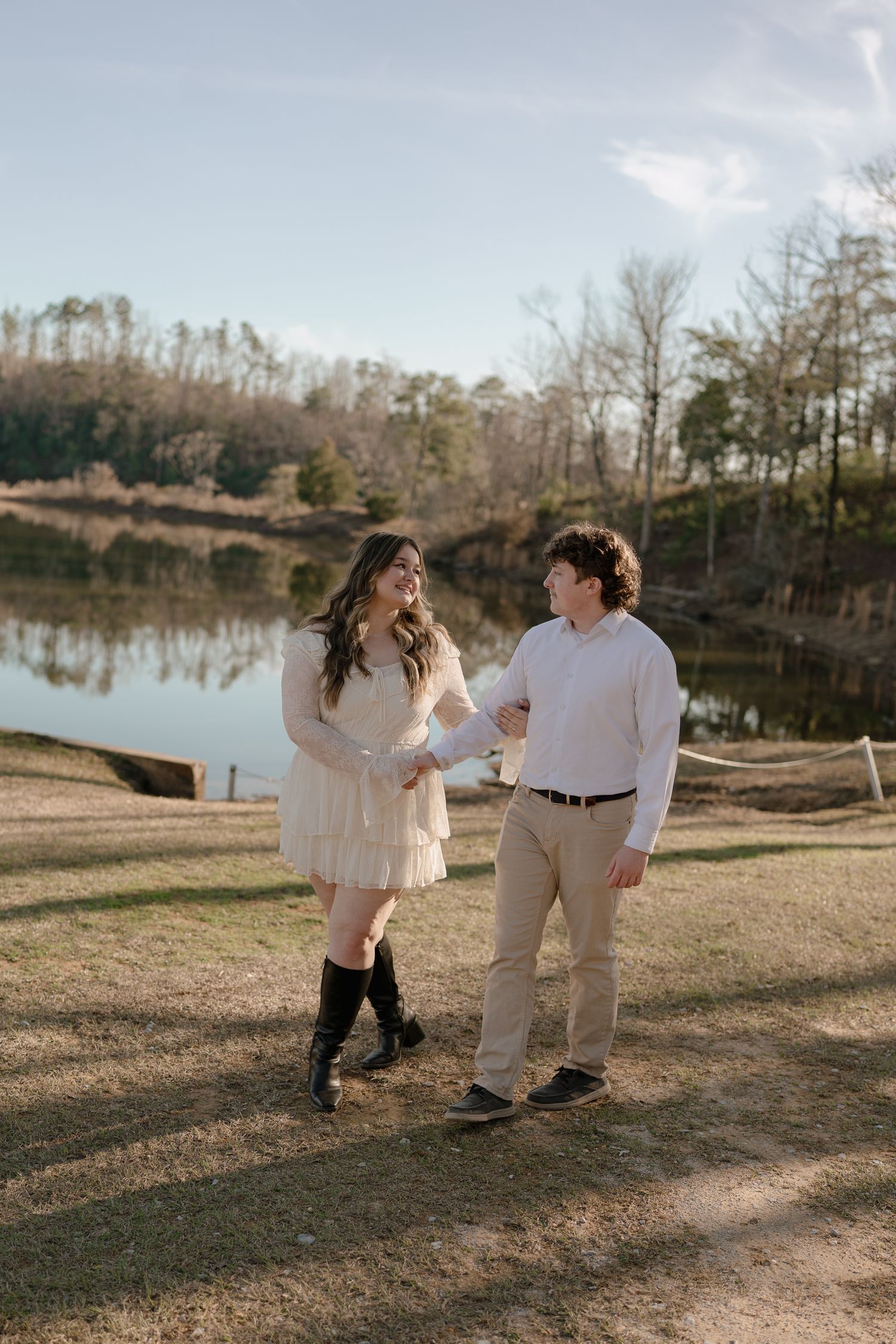 Couple holding hands and smiling at each other by a peaceful lake during golden hour.