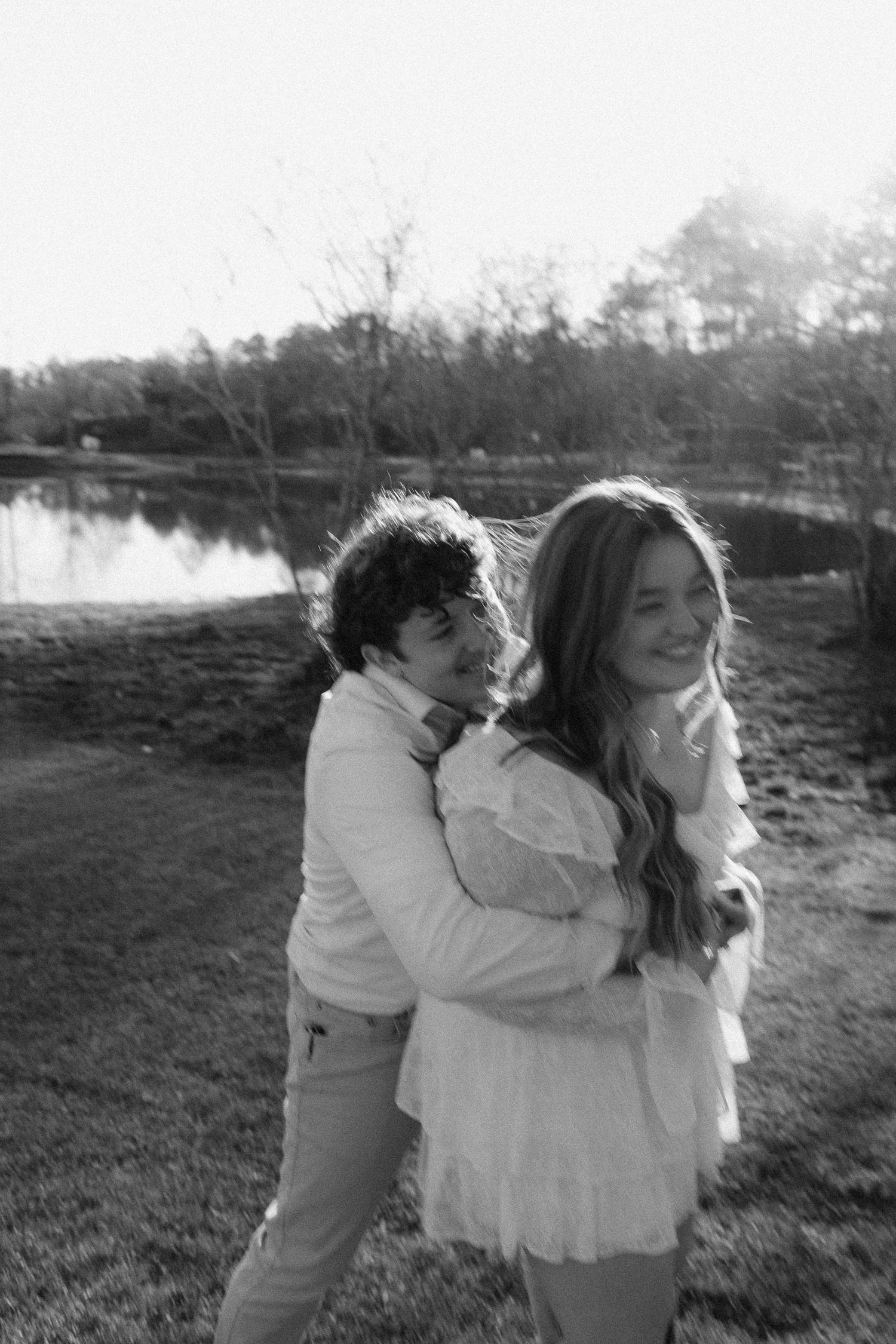 Black and white photo of man embracing woman from behind near lake as she smiles joyfully.