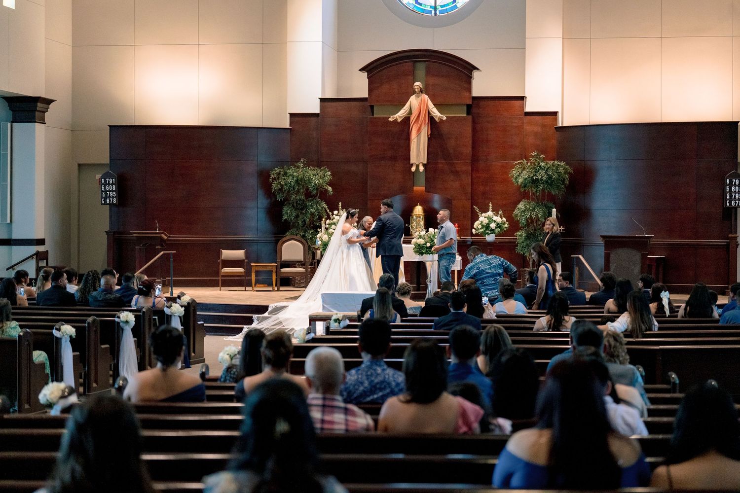 A wedding ceremony takes place in a church sanctuary with wooden pews and a large crucifix mounted on the wall.