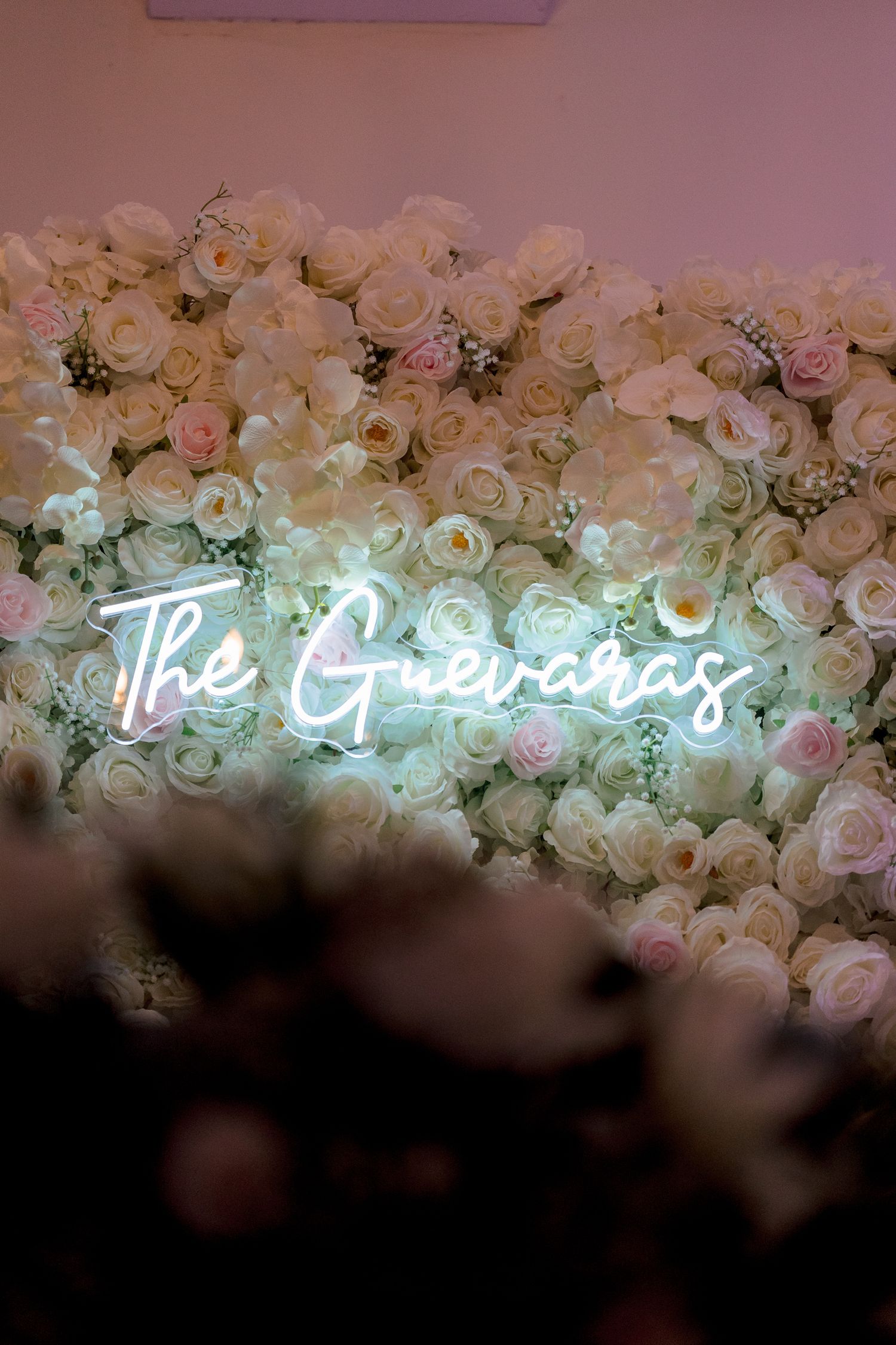 Neon sign reading 'The Gardens' mounted on a wall decorated with pink and white roses creating a romantic ambiance.