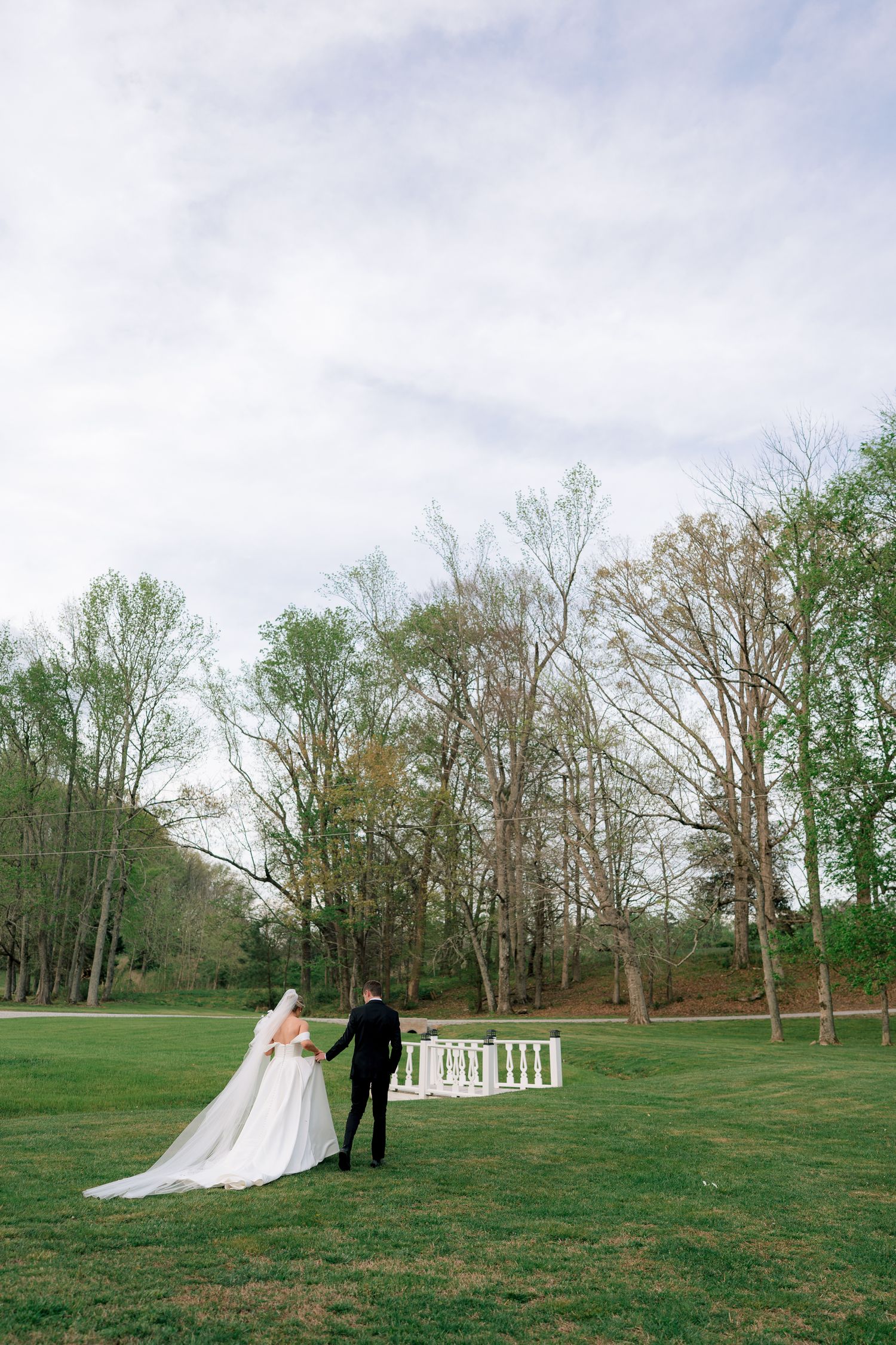 Bride and groom walk hand in hand across lush green lawn toward white garden fence surrounded by tall trees.