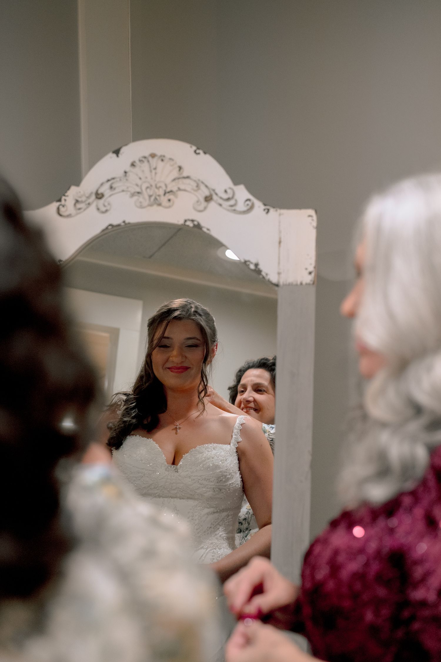 Bride in lace wedding gown smiling in ornate white mirror while mother helps with final preparations.