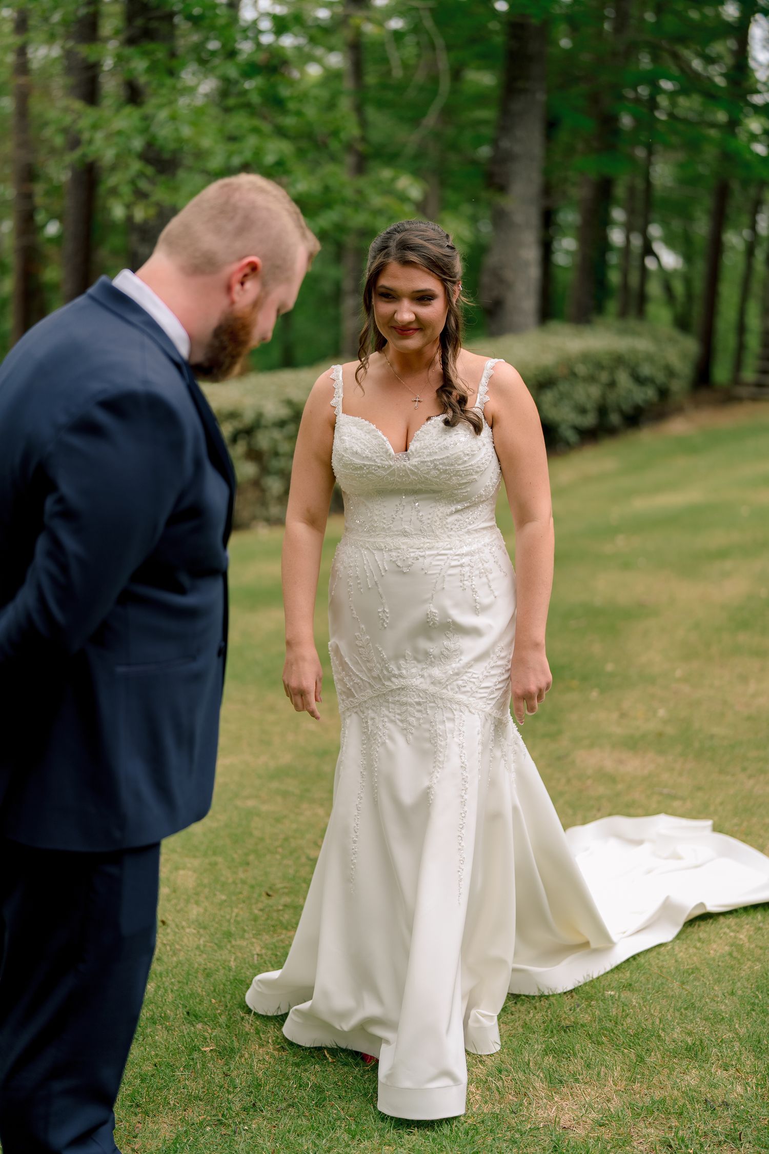 Bride in fitted lace wedding gown smiles at groom during outdoor first look on lush green grass.