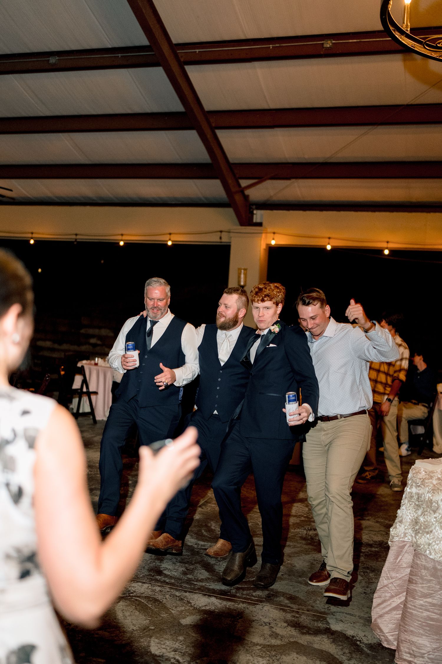 Groom and groomsmen dance together in a line at indoor wedding reception under string lights.
